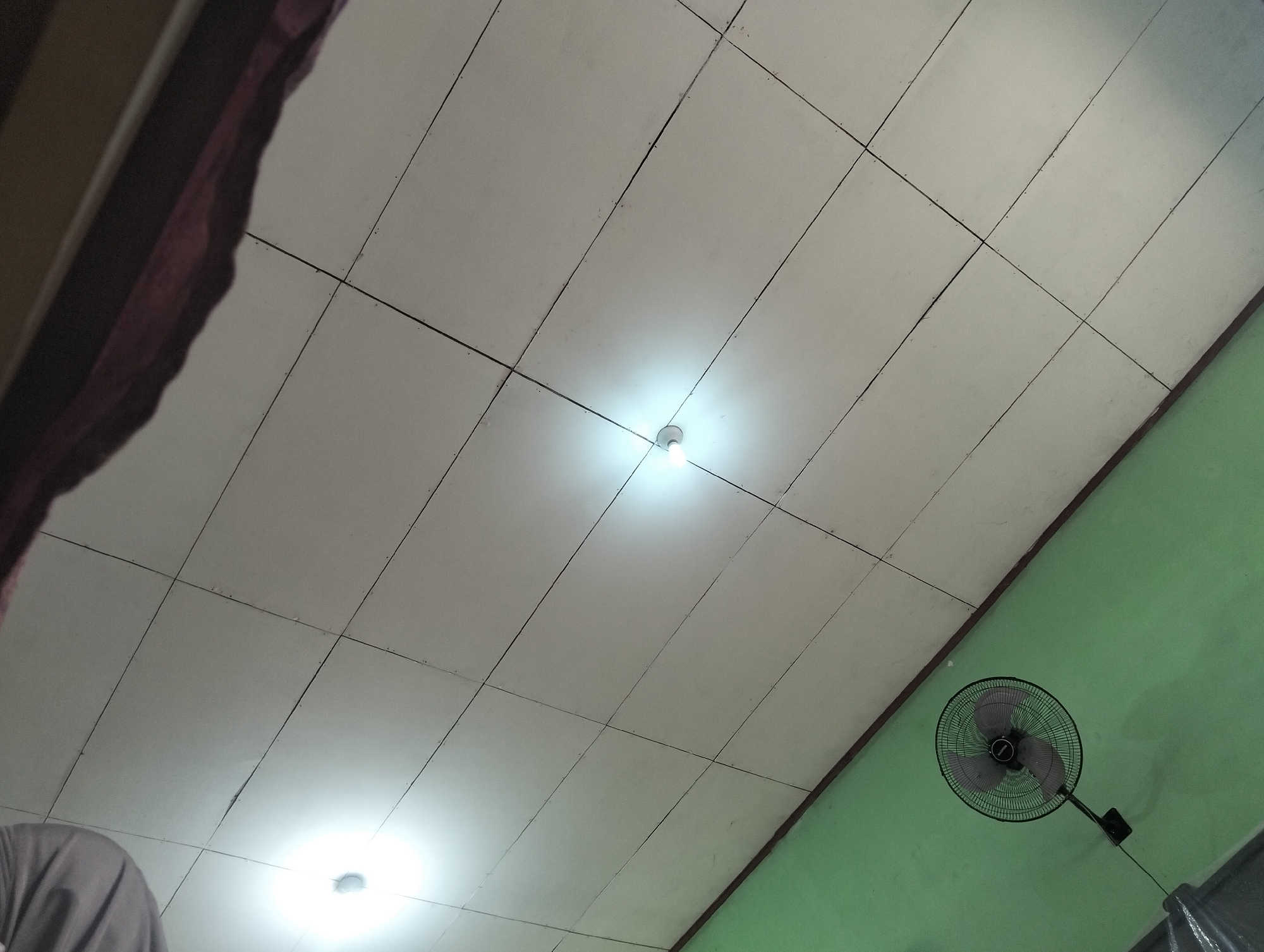 The image captures an interior scene, looking upwards towards a white tiled ceiling. A single bare light bulb glows brightly near the center of the ceiling, casting a luminous halo. To the right, a portion of a pale green wall is visible, with a black oscillating fan mounted on it. The fan is angled, with its blades partially obscured by the grille. In the upper left of the frame, a dark, draped fabric, possibly a curtain or part of a bedsheet, hangs down. The overall impression is of a simple, functional room.
This scene is located in Banjar Agung, Indonesia.