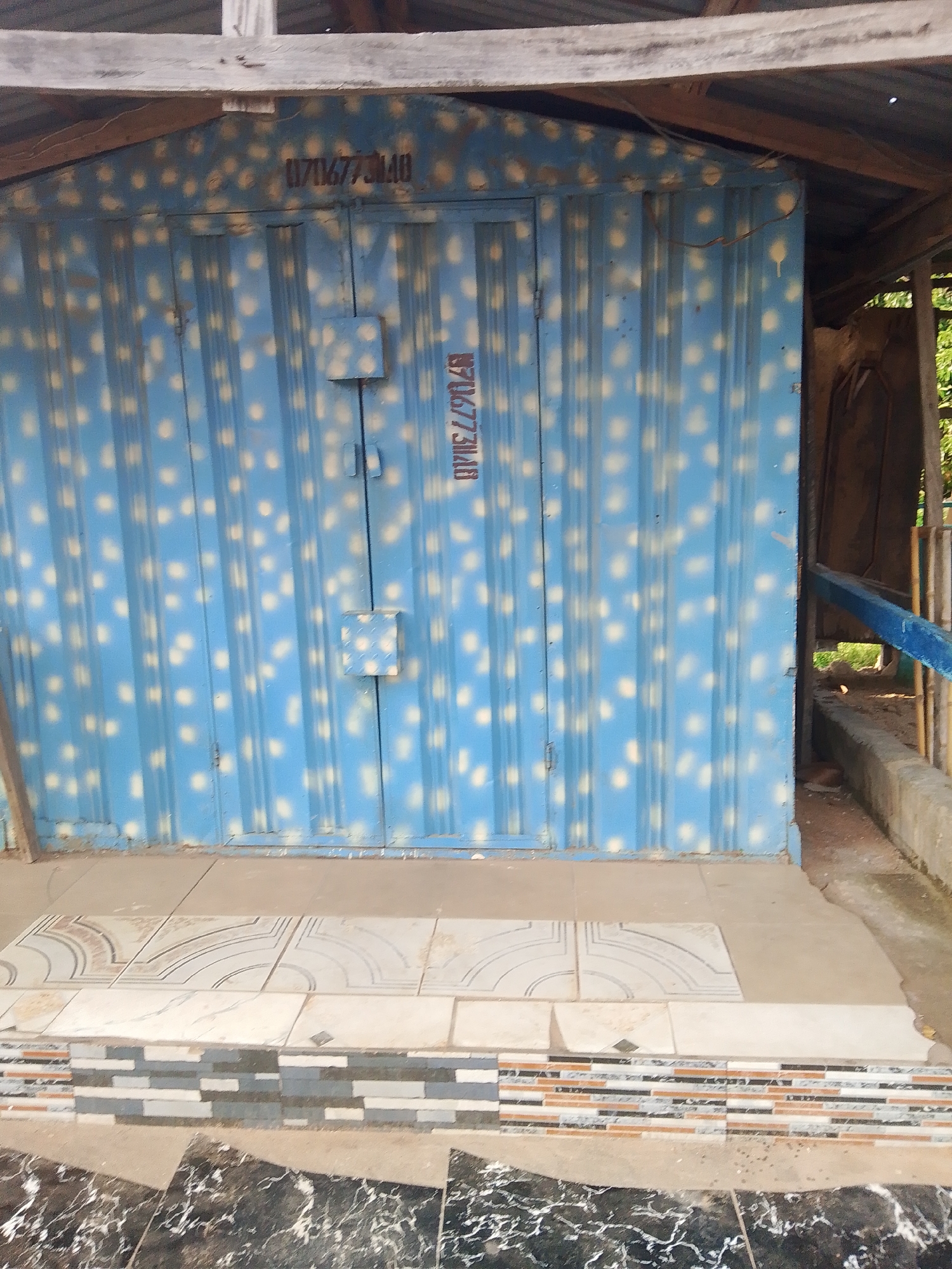 The image shows the blue corrugated metal doors of a small structure, possibly a kiosk or storage unit, located in Jalingo, Nigeria. The doors are spray-painted with a pattern of light-colored dots. A serial number, "011716773140", is stenciled in dark brown paint above the doors. Another stenciled number, "1171677314B", is located vertically on the right side of the doors. The structure appears to be partially covered by a wooden roof with a corrugated metal covering. The foreground shows a tiled step leading up to the structure. The area around the structure appears to be an outdoor environment with some concrete elements and vegetation visible in the background.
