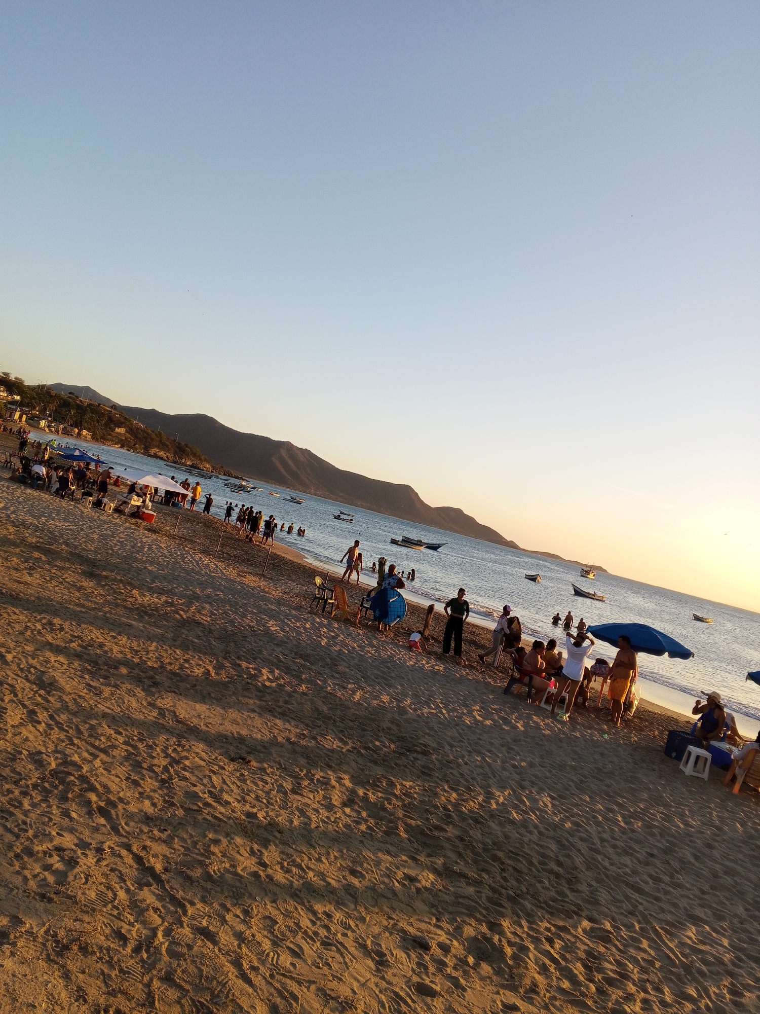 The image depicts a beach scene at sunset. The location is identified as Juan Griego, Venezuela.
People are gathered along the shoreline; some are standing, sitting, or walking, and some are in the water.
Several small boats are anchored offshore.
Umbrellas and tents provide shade on the sand.
Mountains are visible in the background. The sky is clear, with a gradient from blue to orange at the horizon.