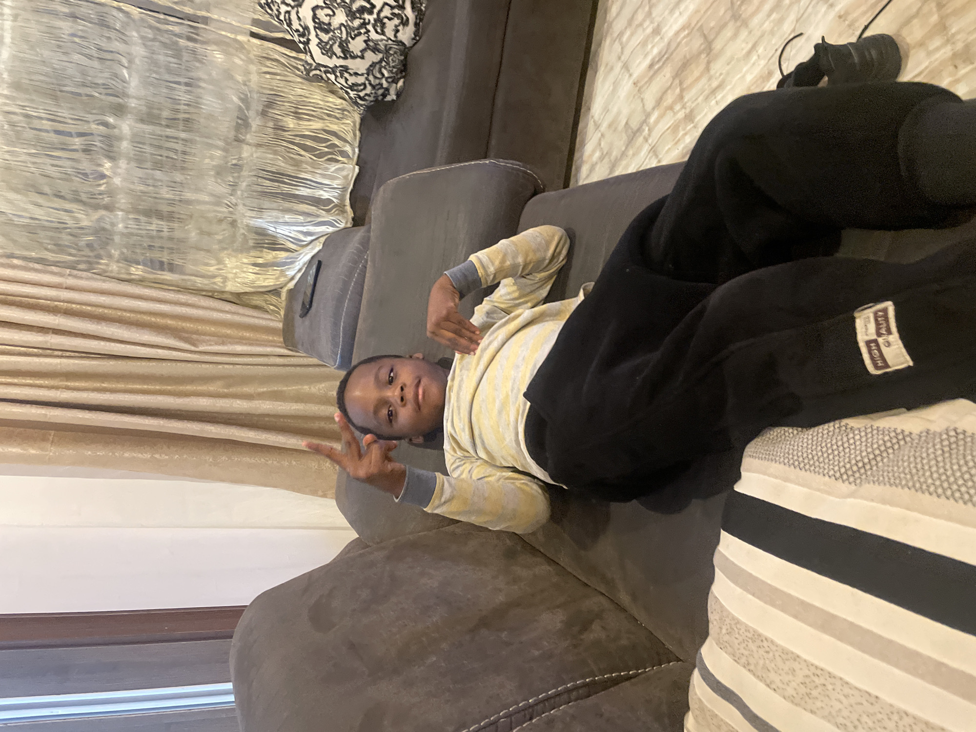A young boy lies on a gray sofa, wearing a long-sleeved yellow and white striped shirt and black pants. He is lying on his left side and gesturing with his left hand, displaying a peace sign. Part of a decorative curtain is visible in the background, and another section of the sofa features a black and white patterned cushion. A black shoe is seen at the far right of the frame. The location is presumably a home setting, but there is no information from the photo that reveals that it is the city of Jos, Nigeria.