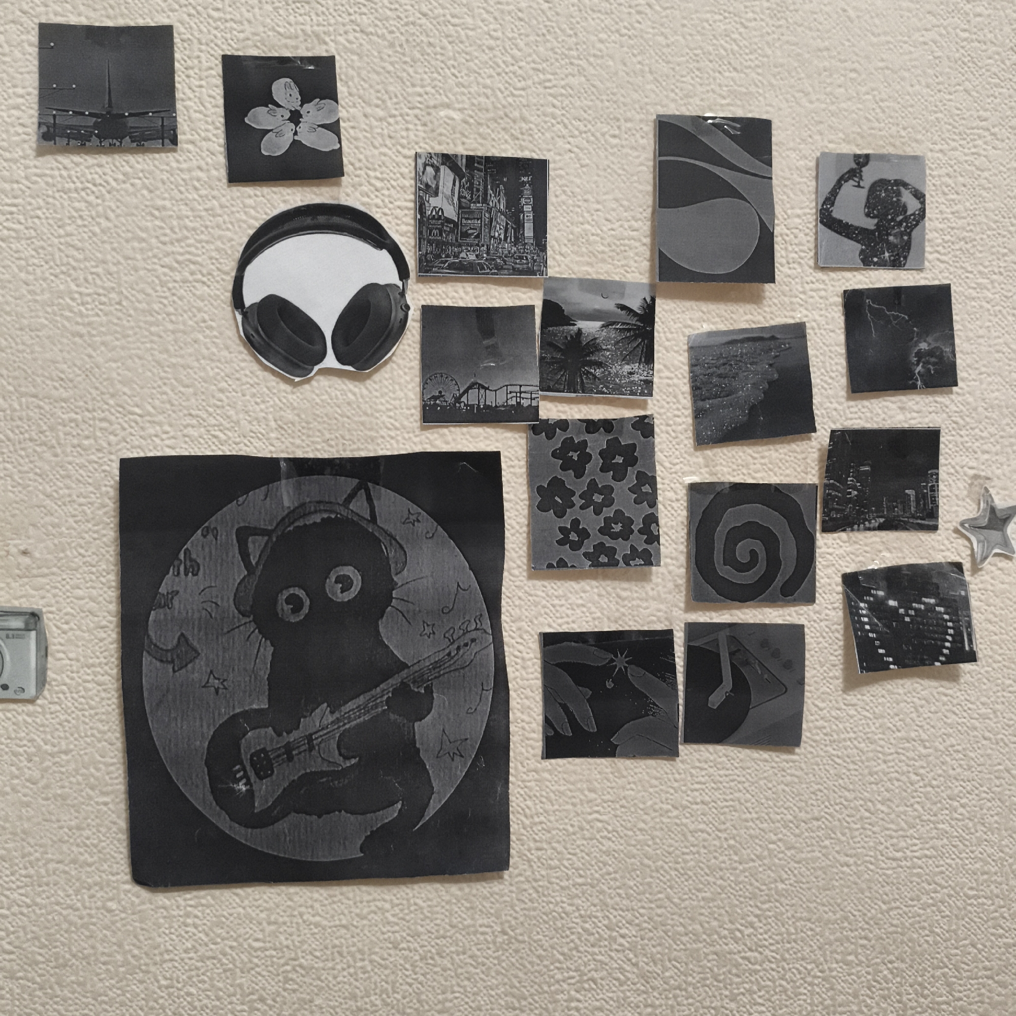 A collection of black and white images are adhered to a textured beige wall. The largest image depicts a cartoon cat wearing headphones and playing an electric guitar, set against a moon backdrop. Other images include an airplane on a runway, a flower with bunny faces on the petals, a pair of headphones, Times Square in New York City, a ferris wheel near a beach, a tropical beach scene, city skylines at night, a person raising a glass, a lightning storm, floral patterns, a spiral design, hand silhouettes, a turntable, and abstract patterns. A small, unidentified object is visible on the lower left side of the image. This image shows a personal, creative arrangement. The location is unknown; the city of Chaikovsky, Russia is not represented in any of the pictures.