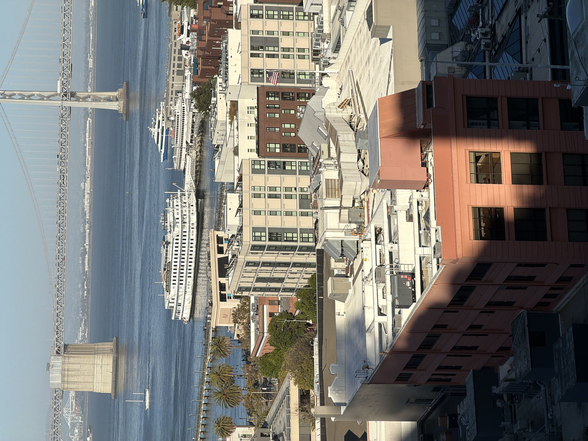 An aerial view of San Francisco, California, United States of America, captures the San Francisco-Oakland Bay Bridge stretching across the water. The bridge's monumental concrete piers and intricate steel superstructure are clearly visible against a clear, blue sky. Several boats, including a white sailboat and a large white paddlewheel boat named "San Francisco Belle," navigate the calm, blue waters of the bay. Along the waterfront, modern buildings with a mix of glass and brick facades are nestled amongst palm trees. The foreground is dominated by rooftops of various buildings, featuring HVAC units, vents, and shadowed architectural details. The lighting suggests it is a sunny day, casting distinct shadows across the urban landscape.