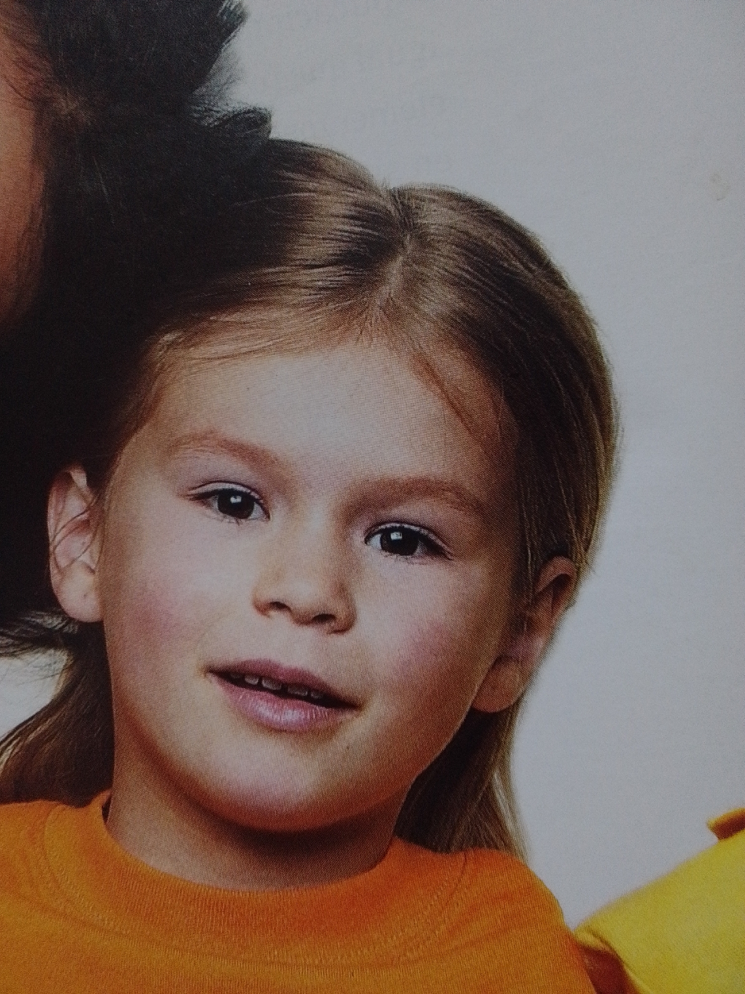 The image shows a close-up of a young girl's face. She has light brown hair and fair skin. Her eyes are dark and looking slightly to the side. She is wearing an orange shirt. The background is a plain, light color. There is no indication of a city or specific location in the image.