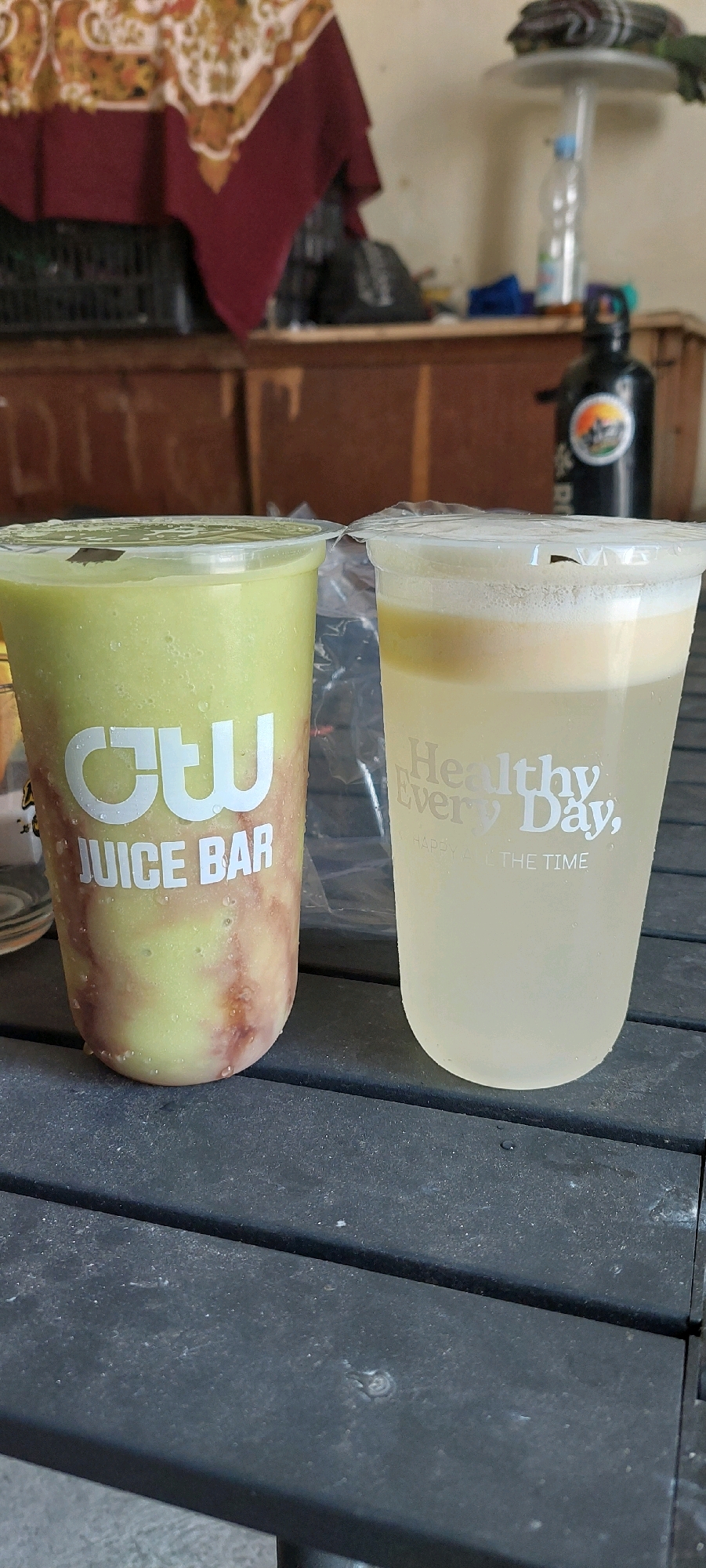 Two clear plastic cups filled with beverages are on a dark, textured surface, possibly a table. The cup on the left contains a thick, pale green liquid with swirls of a reddish-brown substance, and white text that reads "CTW JUICE BAR". The cup on the right contains a lighter-colored liquid with a foamy top layer, and white text that reads "Healthy Every Day," followed by smaller text reading "HAPPY ALL THE TIME." In the background, out of focus, are wooden surfaces, a dark object, and a black bottle with a circular logo. The lighting suggests an indoor or shaded outdoor setting.