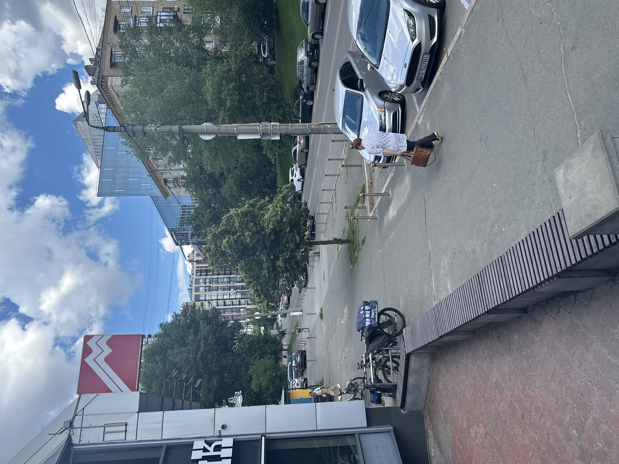 The image captures a street scene in Kyiv, Ukraine, under a cloudy sky, suggesting it could be daytime with variable weather. In the foreground, a person is seated on a bench, holding a brown bag and looking downwards. Beside them are parked bicycles, one with blue delivery bags bearing the text "Domino's Pizza". The street is lined with cars parked on one side, and trees and buildings on the other.
In the background, a modern glass-fronted building stands adjacent to a more traditional brick building with balconies. Further down the street, more apartment buildings and trees are visible. A prominent red sign with a white zig-zag design is mounted on top of a building on the left side of the frame. The overall impression is a typical urban environment with a mix of old and new architecture, indicating a bustling city life. The presence of parked cars, bicycles, and a pizza delivery setup suggests an active street with commerce and daily activities.