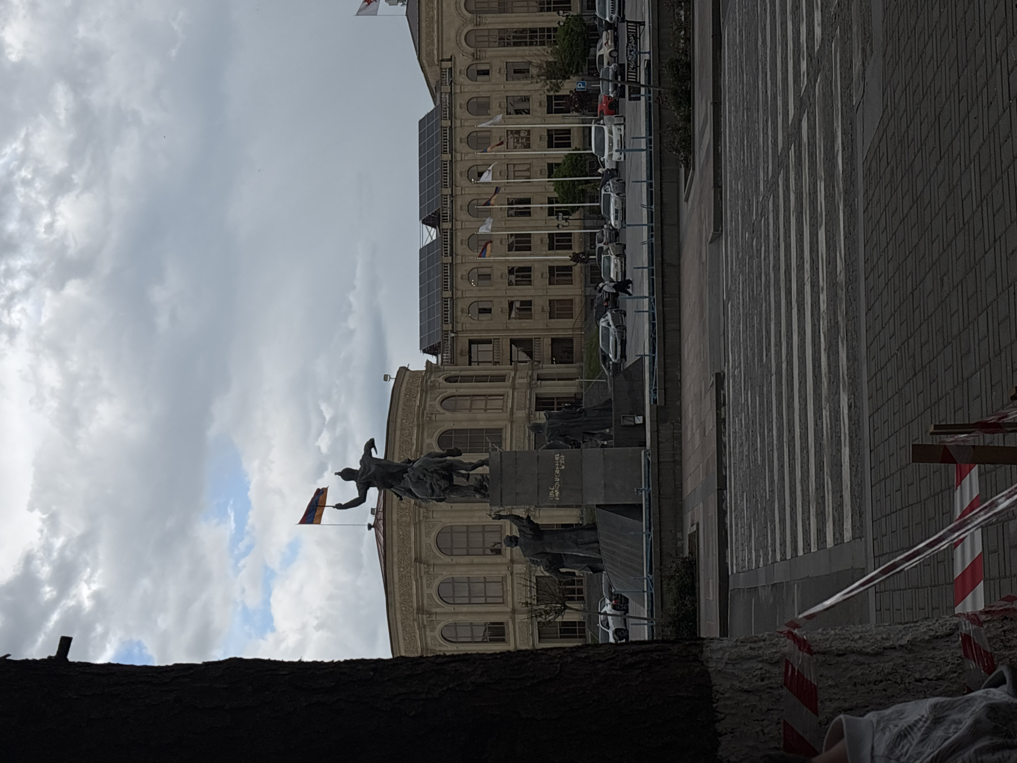 The image shows a wide shot of a public square in Gyumri, Armenia. In the foreground, a tree trunk is visible on the left side, with red and white caution tape wrapped around its base and extending towards the right. A pedestrian crossing is present in the middle ground, with cars parked along the right side of the square and in front of buildings. The background features a large, ornate, light-colored building with arched windows and a dome-like structure. In front of this building, a monument with several statues is erected. One statue depicts a person on horseback holding an Armenian flag. Several other flags are displayed on poles in front of the building. The sky is overcast with clouds.