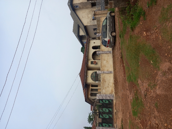 The image shows a residential area in Ita Oniyan, Nigeria, located at latitude 7.293 and longitude 5.169. There is a single-story house with a brown roof and a beige facade. The house has a large, open-air porch with a decorative archway. There is a grey minivan parked in front of the house. The car is facing the street, and there are several power lines running across the sky. The house has a black gate with green panels on the front. The front yard has a patch of grass, and the rest is dirt. There are no people present in the image. The image was taken during the day, and the sky is clear and blue.