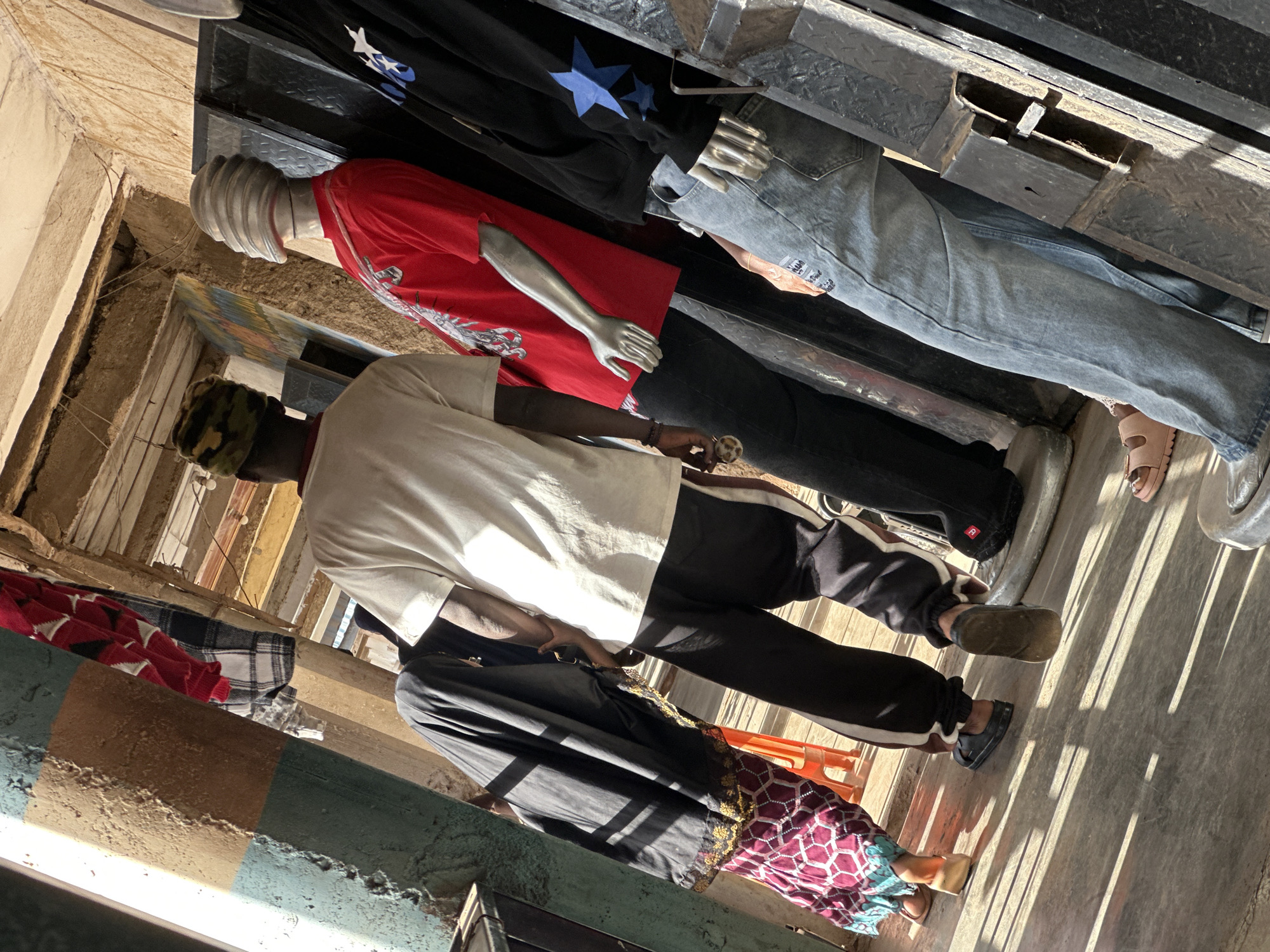 The image shows a street scene in Bukuru, Nigeria. There are several mannequins dressed in clothing, along with what appear to be people. The mannequins are wearing a red t-shirt, a black shirt with blue stars, and jeans. One mannequin is wearing a white t-shirt and black pants. There is also a person wearing a black dress with a patterned skirt and sandals. The scene appears to be a marketplace or a clothing store. The lighting suggests it is daytime, with strong shadows cast by the sun, indicating a bright, sunny day. The overall impression is that of a busy commercial area, likely outdoors. The ground is concrete, and there are wooden and concrete structures visible. The walls are rough and textured. Some of the clothing on the mannequins and the person are brightly colored. The scene is tilted, suggesting it was taken from an angled perspective.
