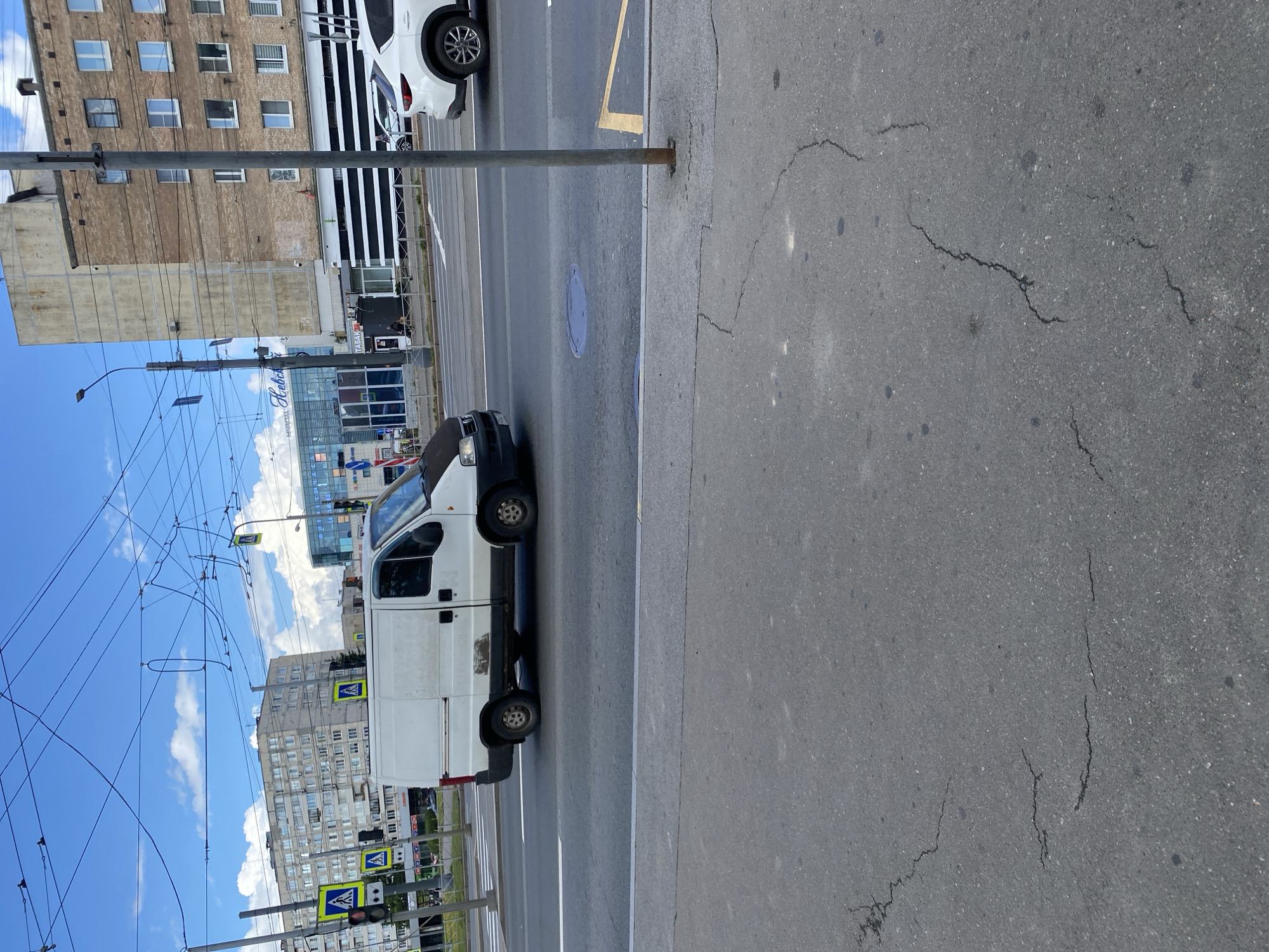 The image captures a daytime urban scene in Saint Petersburg, Russia, likely near the intersection of Prospekt V. I. Lenina and Botanicheskaya Street, given the presence of a Metro station sign ("Botanicheskaya"). A white utility van with a dark hood is driving on the asphalt road, which has visible cracks and patches. In the background, several multi-story buildings are visible, some appearing to be residential blocks with numerous windows, and one with a prominent "Nevsky Center" sign, indicating a commercial area. Overhead, a complex network of trolleybus wires stretches across the sky, with a blue sky and scattered clouds above. Pedestrian crossing signs with figures are visible on traffic light poles on the left side of the frame. A white SUV is parked or driving on the road to the right of the van. A metal pole stands vertically on the right side of the image, along with a yellow painted line on the road, possibly indicating a parking or no-stopping zone. The foreground is dominated by the cracked surface of a sidewalk or pedestrian area, also made of asphalt.