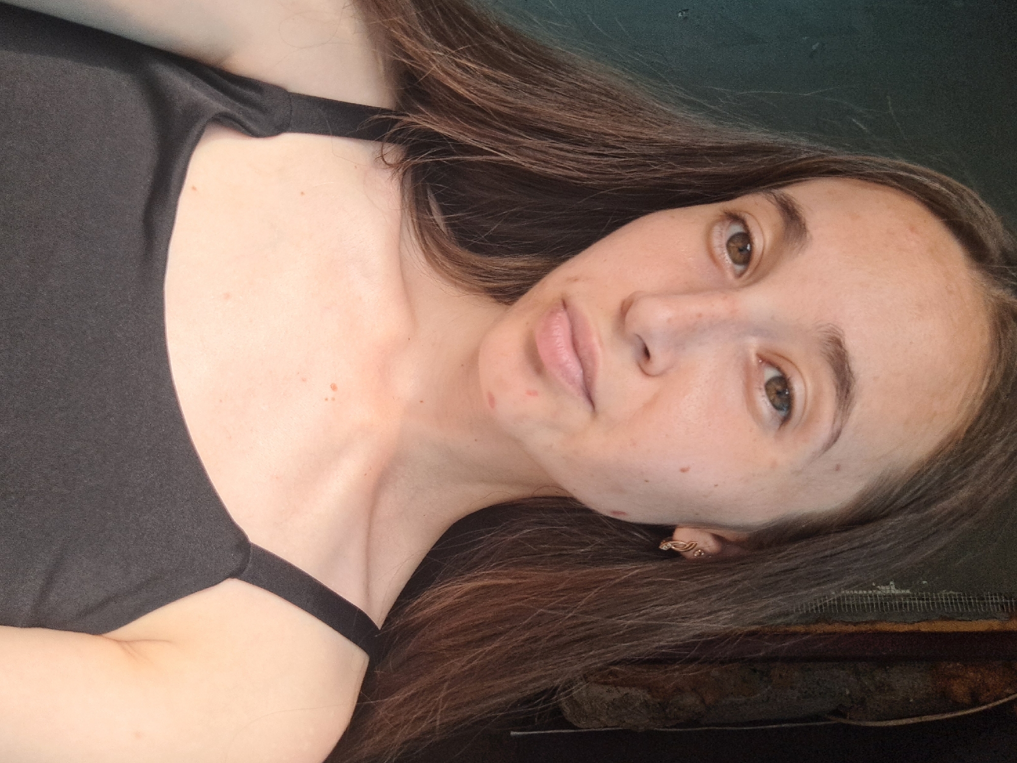 This is a close-up selfie of a young woman with long, dark brown hair, taken outdoors. She is wearing a black tank top. She has brown eyes and a light complexion with some blemishes and freckles. She is wearing a gold earring on her right ear. The background appears to be a wall with a dark green texture on the left and a darker, rougher textured area on the right, possibly a doorway or building facade. There is no indication of the specific location within Vypasne, Ukraine.