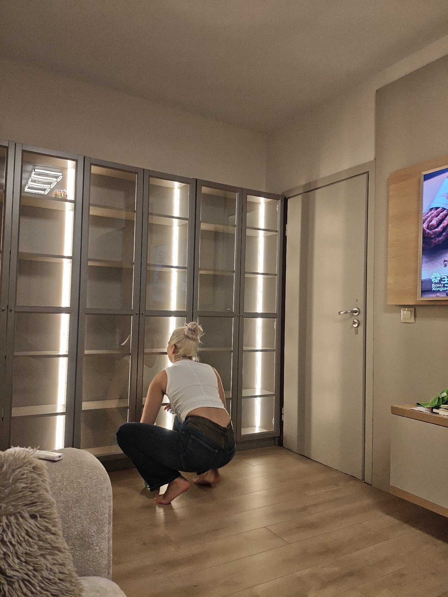 A woman is crouching on a wooden floor in a room. She is facing a tall, gray shelving unit with glass doors and built-in vertical lighting. She has blonde hair styled in a bun and wears a white sleeveless top and blue jeans. The floor is made of light brown wooden planks. To the right of the shelving unit is a closed gray door with a silver handle and a key inserted in the lock. To the right of the door, there is a wall-mounted flat-screen television above a console table. The location and city are not evident in the image.