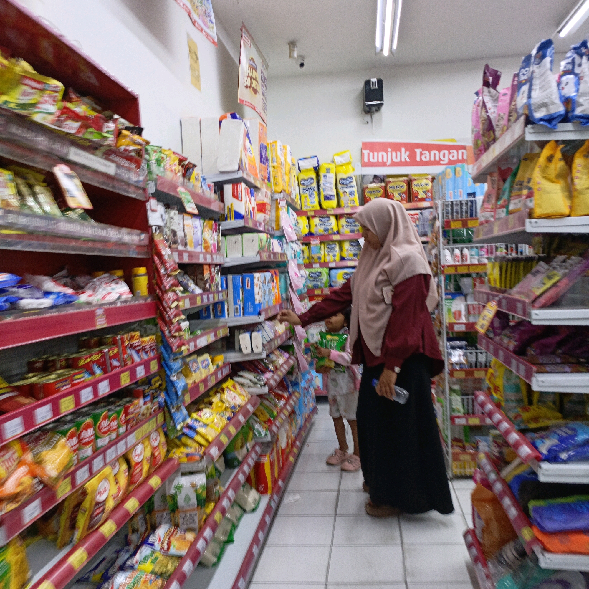 The image shows an interior view of a brightly lit convenience store, likely a minimarket, located in Salatiga, Indonesia. Rows of shelves are stocked with various packaged goods, including food items like instant noodles, snacks, and canned goods, as well as household products such as diapers and detergent. The shelves are red with white price tag holders.
In the foreground, a woman wearing a light pink hijab and a maroon long-sleeved shirt is seen from the side and from the back. She is holding a clear plastic bottle of water in her right hand and appears to be reaching for an item on a shelf with her left hand. Standing next to her, slightly in front, is a young child wearing a pink and white patterned outfit and pink shoes. The child is looking at the shelves and holding a green packaged item.
In the background, more shelves are visible, filled with merchandise. A red sign with white text reading "Tunjuk Tangan" (meaning "Point Hand" in Indonesian) is displayed prominently on the right side. The floor is tiled with white and light grey squares. The overall impression is a typical retail environment in Indonesia.