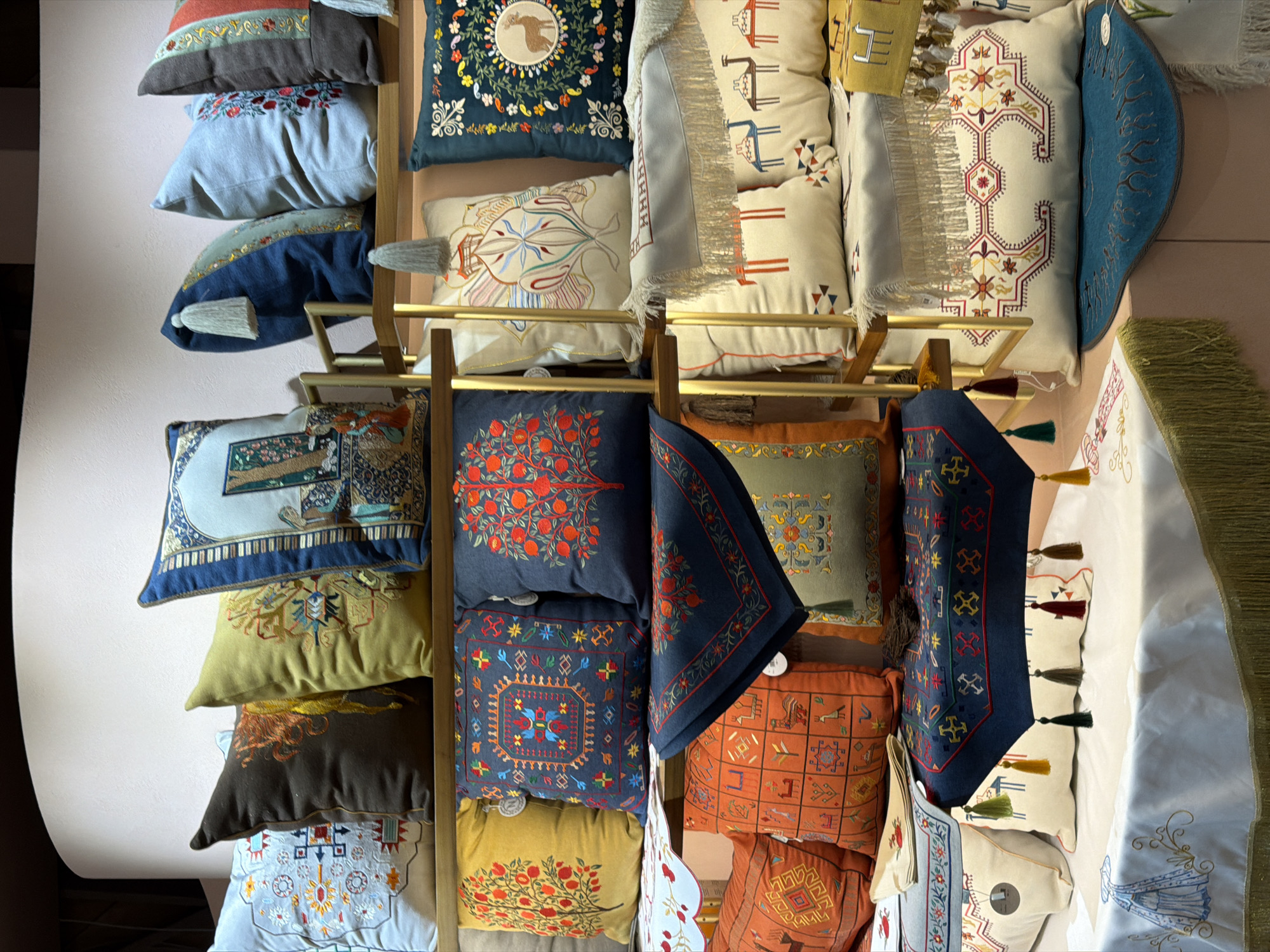 The image displays a retail display of decorative throw pillows and textiles. The items are arranged on multiple shelves, with a focus on intricate embroidery and geometric patterns. The pillows feature various designs including stylized trees, animal motifs, and traditional ethnic patterns. Some pillows have tassels and fringe details. The textiles are laid out as runners or accent pieces. The backdrop appears to be a wall with a curved top. The display is well-lit, highlighting the textures and colors of the products. The location is specified as Khazar Raion, Azerbaijan.