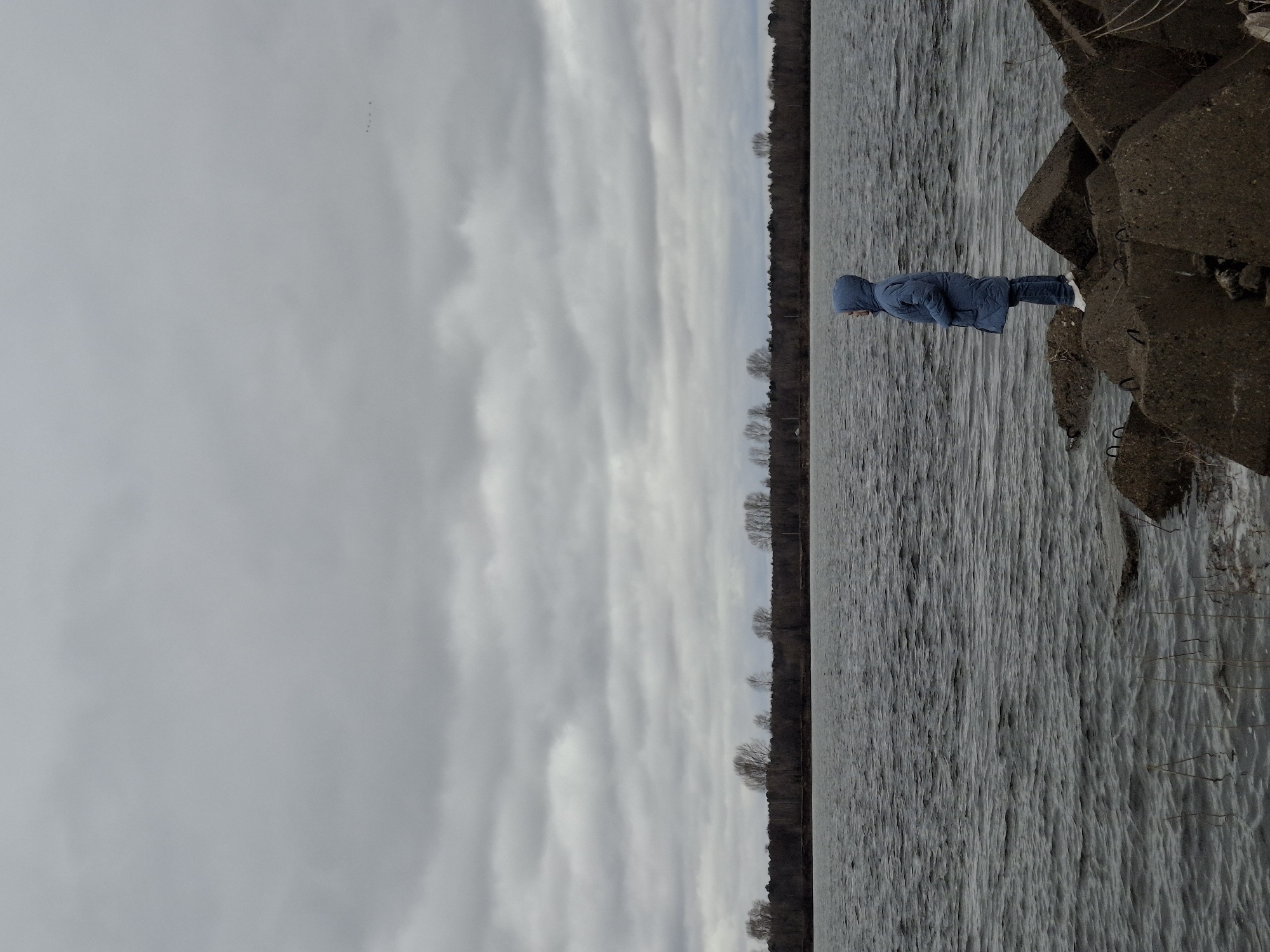 A person in a blue hooded coat stands on large concrete blocks next to a body of water, likely a lake or river, under an overcast sky. The water's surface is disturbed by ripples. A thin line of trees is visible on the far side of the water. The location is inferred to be in Chaikovsky, Russia based on image metadata.