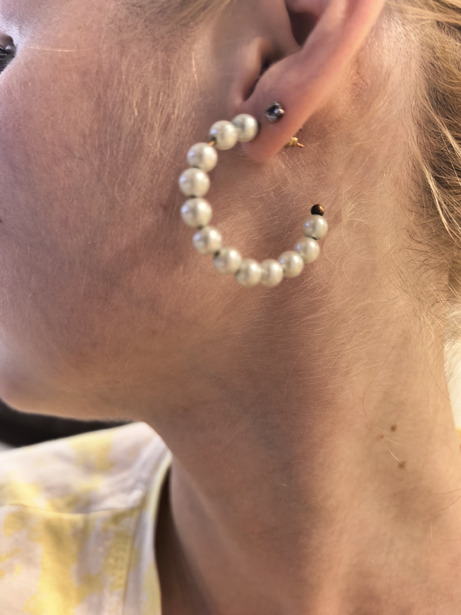 The image shows a close-up, side profile of a person's ear and neck. The person is wearing a large hoop earring adorned with white beads and a smaller stud earring in the lobe. A portion of their hair, which appears blonde, is visible. The person is wearing a yellow and white patterned garment, likely a shirt. The background is blurred and shows a neutral, indoor setting, with no discernible landmarks to indicate a specific location in Sterlitamak, Russia. The focus is on the jewelry and the side of the person's face and neck.