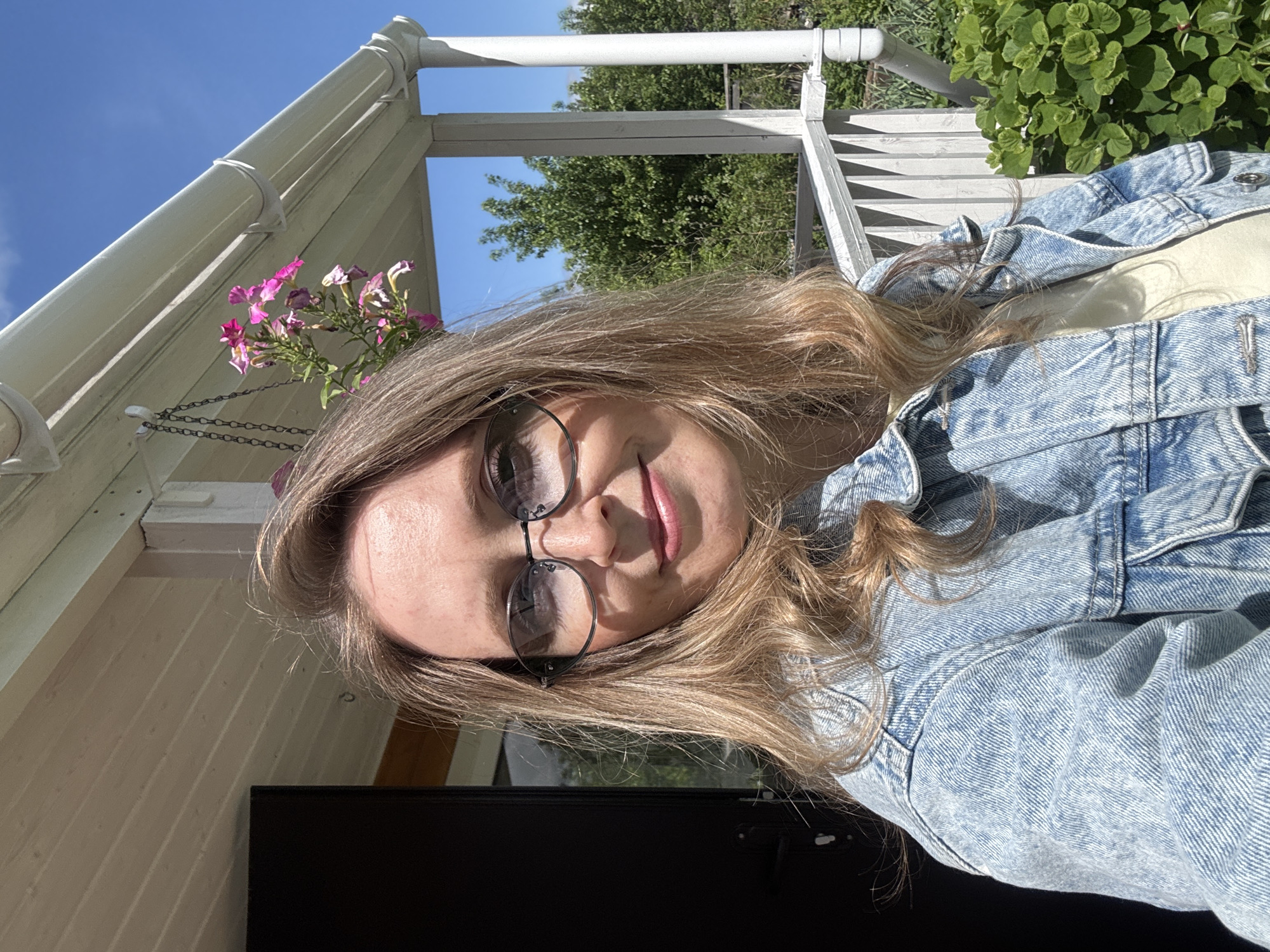 A woman with long, wavy blonde hair and wearing round-framed glasses is taking a selfie. She is wearing a light-wash denim jacket. Behind her is a white wooden structure that appears to be a porch or part of a building. There are hanging flowers to her left and greenery behind her. The sky is blue, suggesting it is daytime. The presence of a black rectangular object partially in the frame at the bottom suggests she may be holding a phone or tablet. No specific identifying details confirm the location as Chaikovsky, Russia.