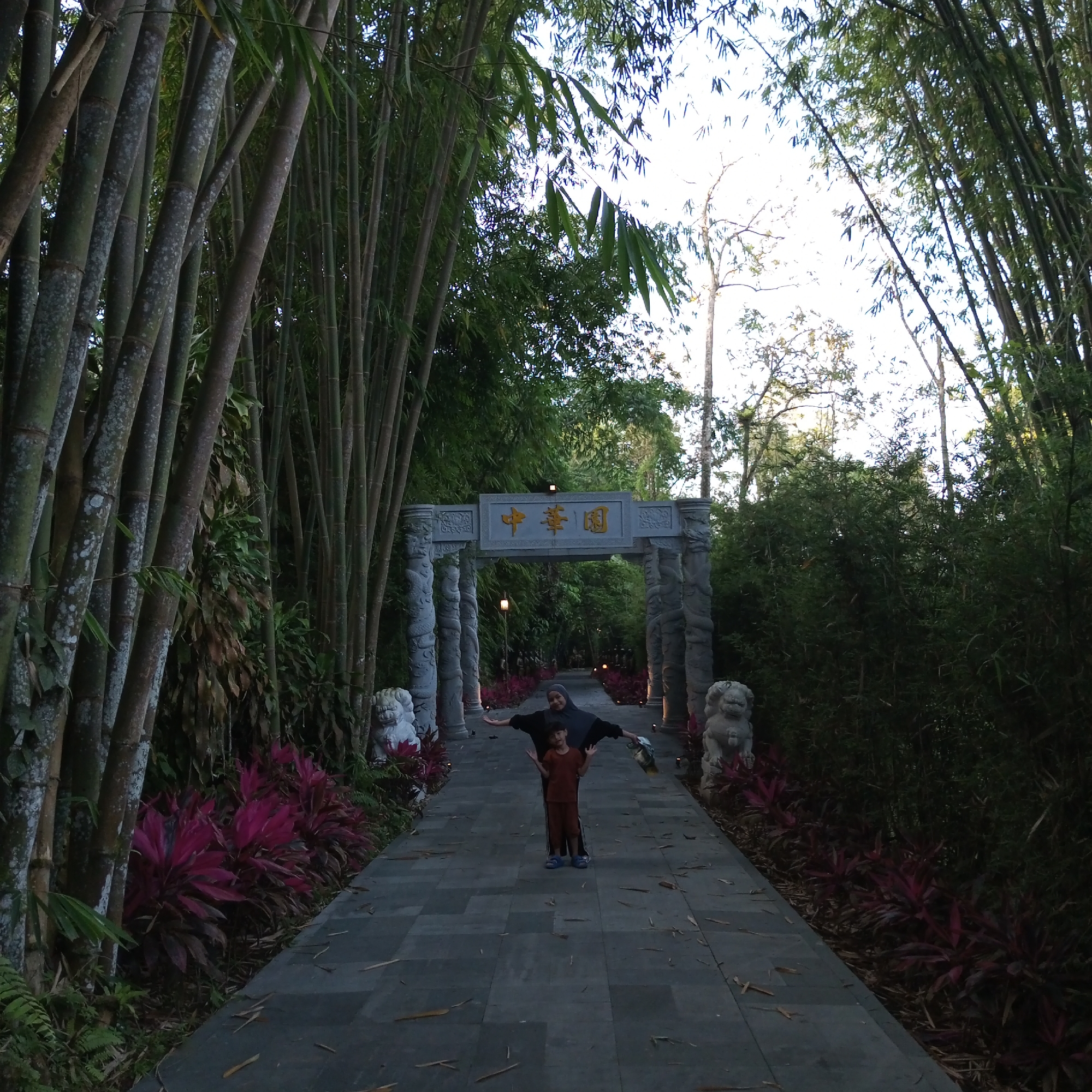 The image depicts a pathway lined with tall bamboo trees on either side. In the foreground, a woman in a dark headscarf and a child in a brown shirt and pants stand on the paved pathway. The pathway leads towards an ornate stone archway with Chinese characters visible on a sign above it. The archway is flanked by two stone lion statues. Red-leafed plants line the sides of the pathway near the archway. The scene is set within an outdoor environment, likely a garden or park, with dense foliage suggesting a tropical or subtropical climate. The lighting indicates it is likely late afternoon or early evening. The location is identified as being in Suka Makmur, Indonesia, though this context is not visually evident within the image itself.