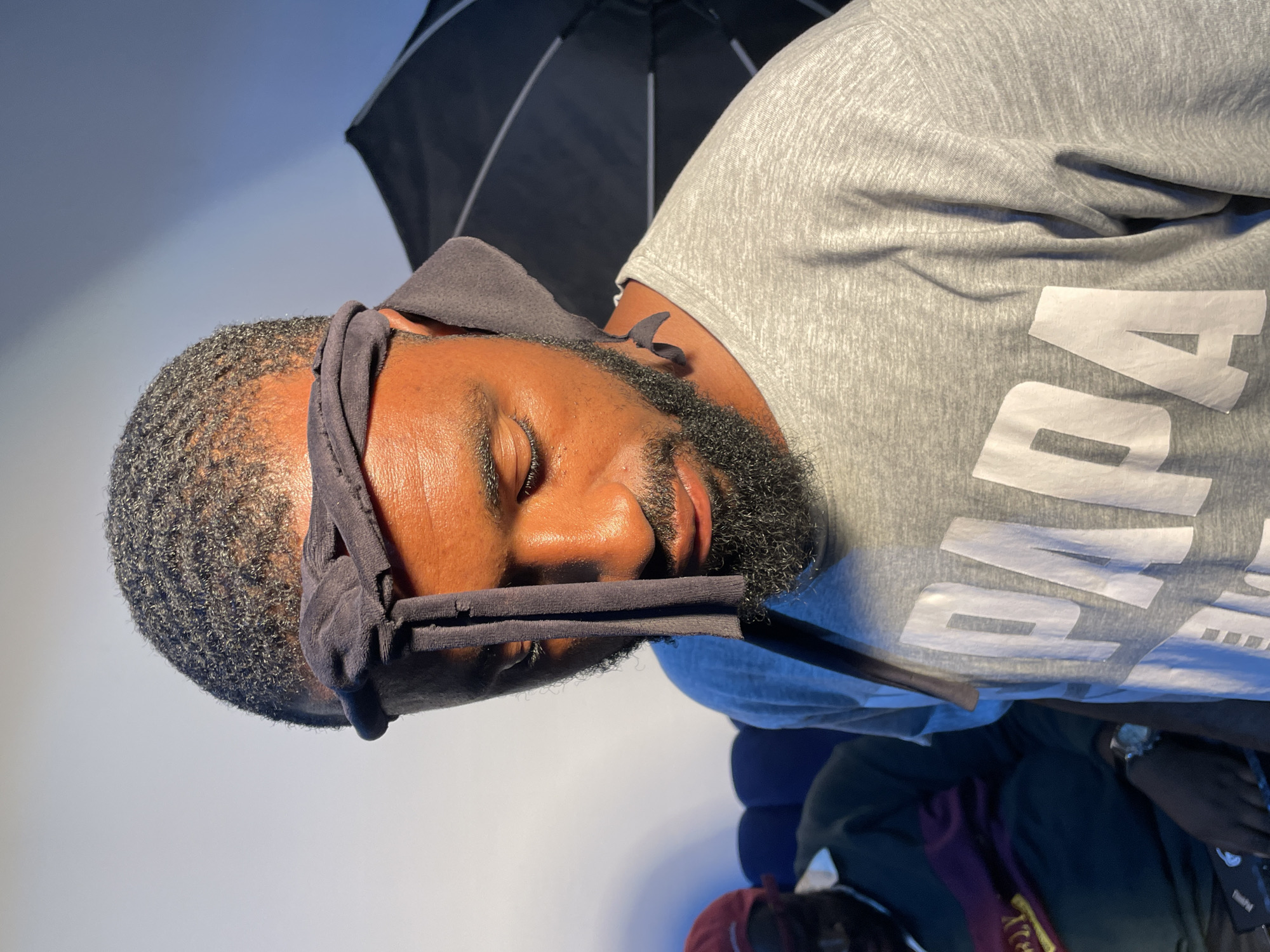 A man with dark skin, a beard, and short, curly hair is lying down with his eyes closed. He is wearing a gray headband and a gray shirt with white lettering on it. A black umbrella is behind his head. He is lying on what appears to be a pile of clothes or blankets. The background is a light blue-gray wall. There is no indication of the location of Barakin Akawo, Nigeria in the image.