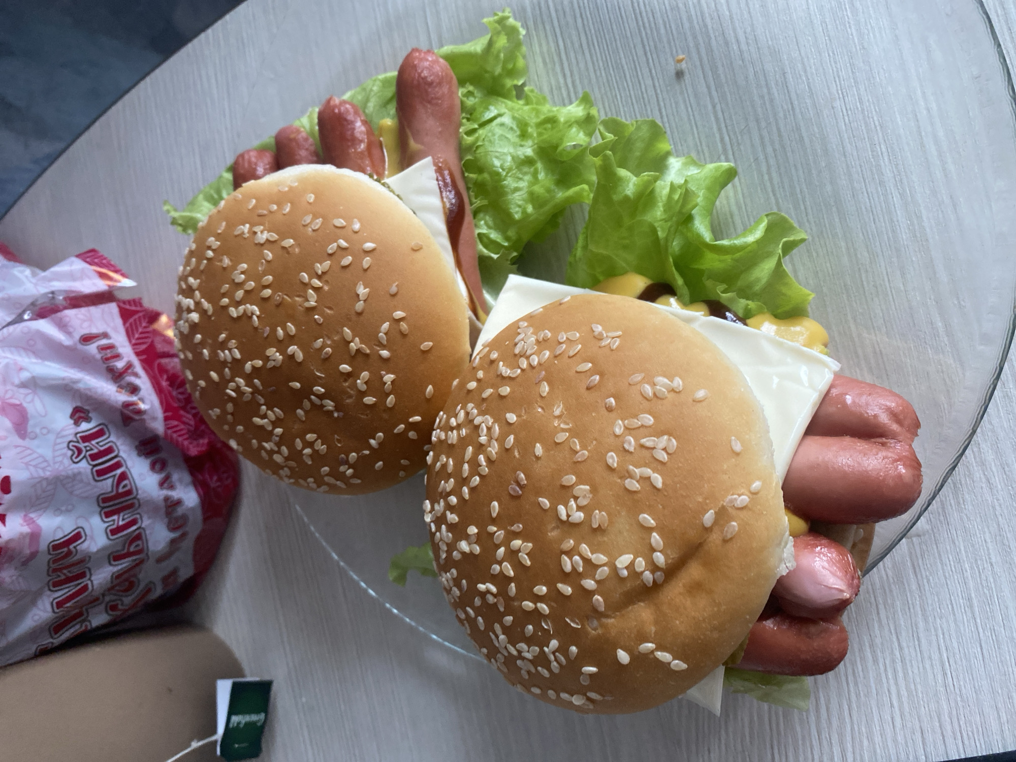 The image shows two hot dogs on a clear plate, likely in a casual setting. The hot dogs are served in sesame seed buns and include cheese, mustard, lettuce, and a sauce, possibly ketchup. A package of some kind of food with Russian text is visible in the background, along with a tea bag on the table. The location is potentially Izhevsk, Russia.