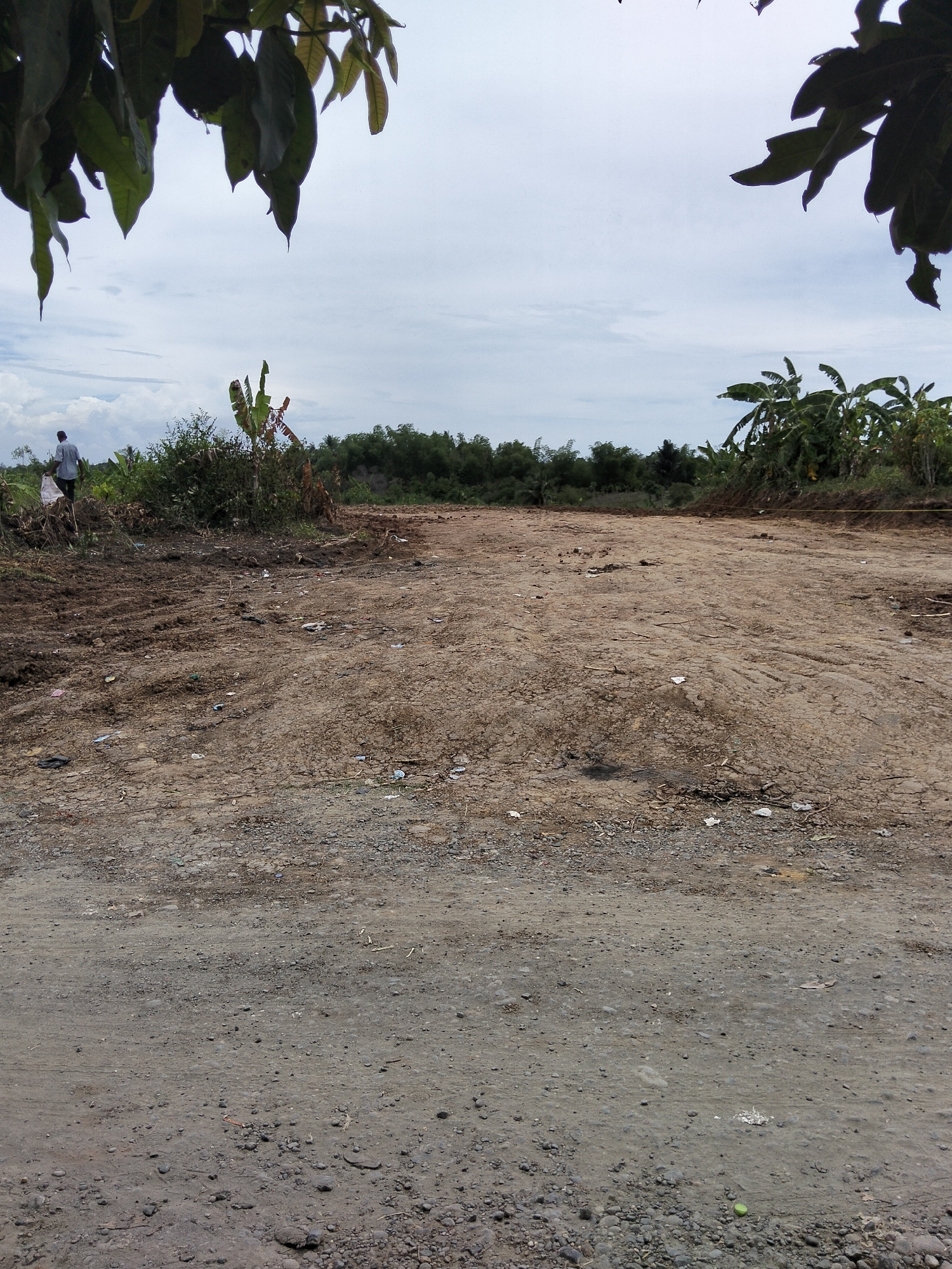 An outdoor, eye-level shot captures a dirt path and cleared land under a cloudy sky in Payagaboh, Indonesia. The path is rough, with loose stones and dirt, leading into a wider, flat area that has been cleared of vegetation. Scattered trash, including plastic bags and wrappers, is visible on the ground. To the left, a person in a dark shirt walks away from the camera, carrying a white bag. Beyond the cleared area, dense greenery and trees form a thick border, with some banana trees prominent on the right. Overhead, the leaves of large trees hang down from the top of the frame. The overall scene suggests a rural or developing area.
