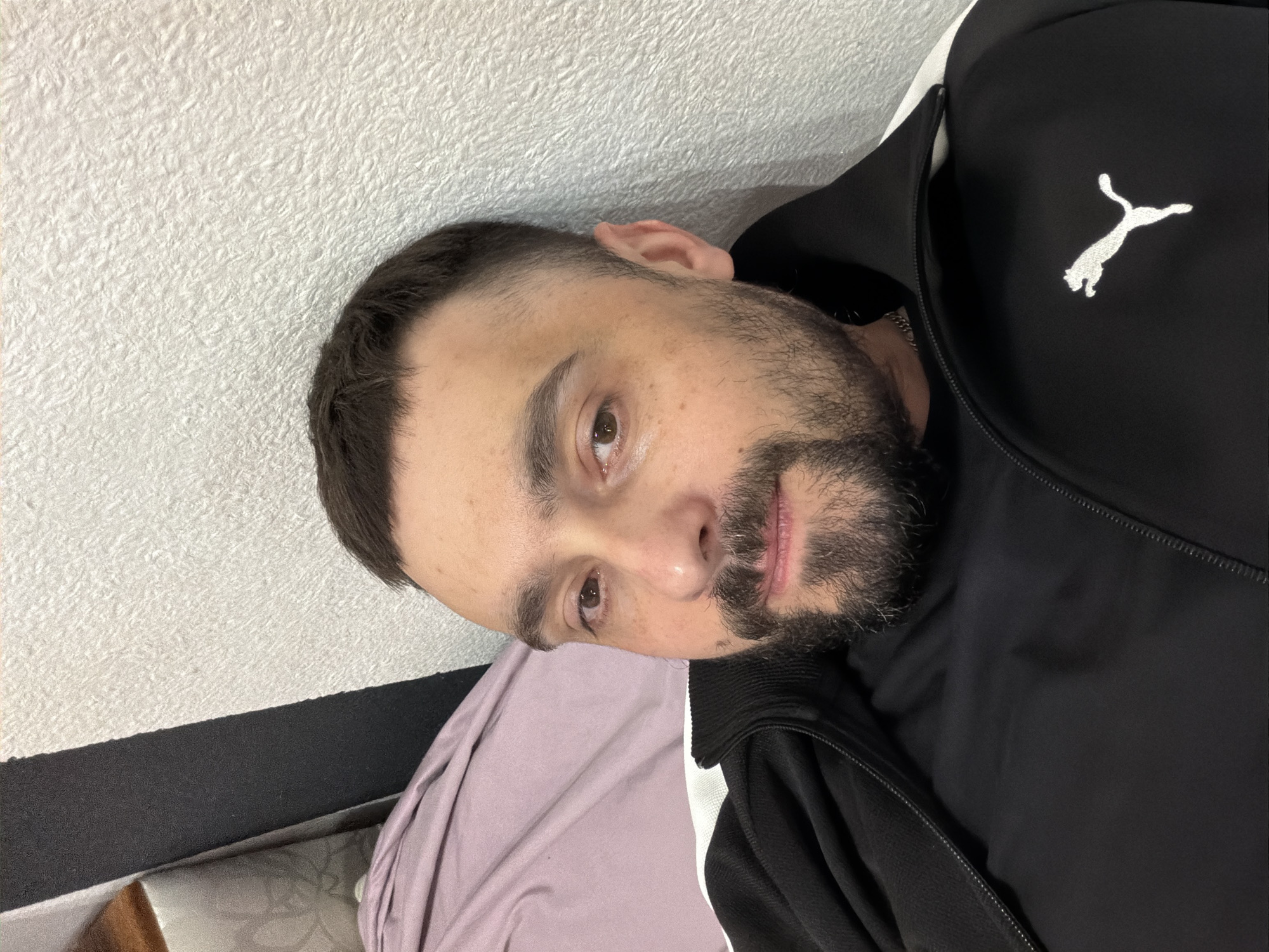 A man with short dark hair and a beard is lying on his side on a bed with a light purple sheet. He is wearing a black Puma track jacket with a white logo on the right chest. The background is a white wall. There is no information in the image to suggest a location in the city of Kovel, Ukraine.