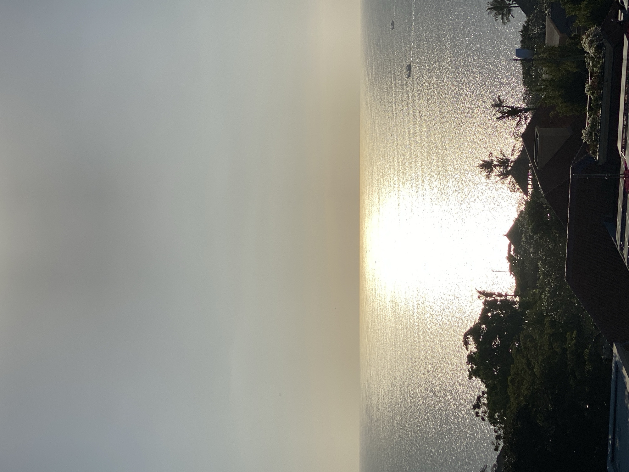 The image shows a wide, calm ocean under a bright, hazy sky. The sun is low on the horizon, creating a strong reflection on the water's surface. In the foreground, there are silhouetted rooftops of buildings and lush green trees, suggesting a coastal or elevated vantage point overlooking the water. A few small boats are visible on the ocean in the distance. The overall atmosphere is serene and tranquil, likely captured during sunrise or sunset.