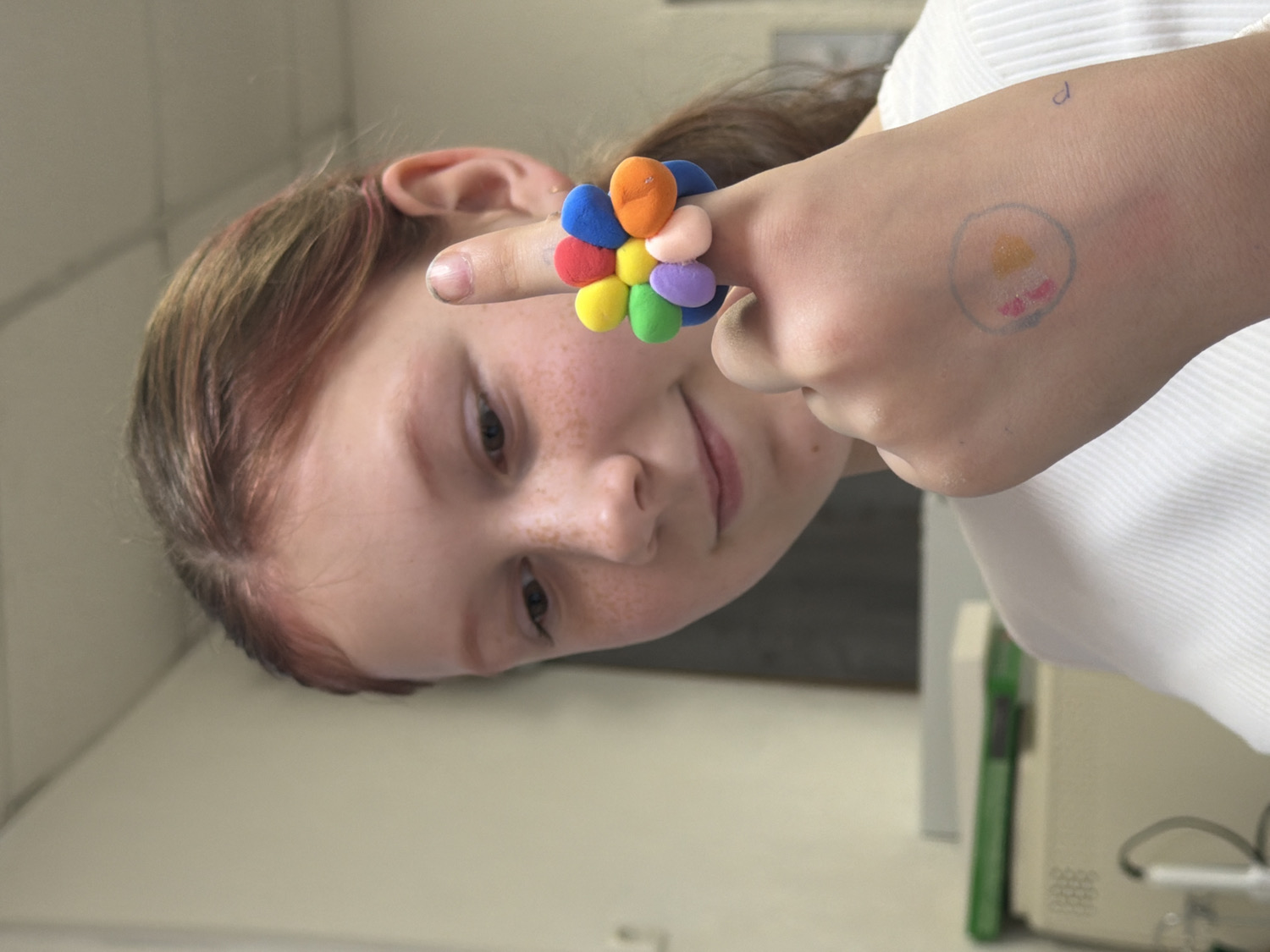 An eye-level, rotated close-up shot captures a young girl indoors in Chaikovsky, Russia, looking towards the viewer with a gentle, soft smile. Her head is tilted, and the image is rotated 90 degrees clockwise. She has light brown hair with reddish highlights, a fair complexion, and numerous freckles across her nose and cheeks. Her dark eyes are open, engaging the viewer.
On her left index finger, she wears a vibrant, flower-shaped ring composed of small, soft, multicolored spheres or beads in shades of orange, yellow, blue, red, green, and lavender. Her fingernails are short and unpolished. Visible on the back of her left hand, near her wrist, is a faded, circular drawing in light blue, containing hints of yellow/orange at the top and pink/red at the bottom, resembling a simple stylized face or sun. Above this drawing, a faint, small handwritten letter, possibly a "d" or similar symbol, is visible.
She is wearing a white, subtly textured shirt. The background is a plain, light-colored wall, slightly out of focus. To the right, blurred beige and green elements of what appear to be office or institutional equipment are discernible, suggesting a potentially non-domestic indoor environment like a school, clinic, or office. The lighting is soft, suggesting an indoor setting without direct sunlight.