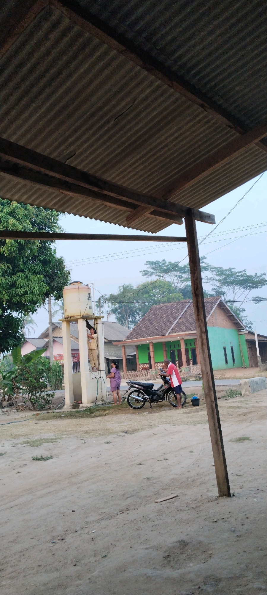 In Tanjung Ratu Ilir, Indonesia, the scene depicts an outdoor area with a sandy ground. In the foreground, a corrugated metal roof supported by wooden beams partially obscures the view. Further back, a white water tank elevated on a concrete structure is visible. A person in a light-colored uniform stands on the platform of the water tank, appearing to be attending to something on its surface. Below the platform, another person in a purple outfit stands next to a black motorcycle. A third individual, wearing a red and white shirt, is also near the motorcycle, seemingly engaged with it. Behind these figures, a green house with a brown tiled roof stands, flanked by other residential structures. Lush green trees and vegetation are scattered throughout the background, suggesting a semi-rural or suburban setting. The sky is hazy, indicating either early morning or late afternoon.
