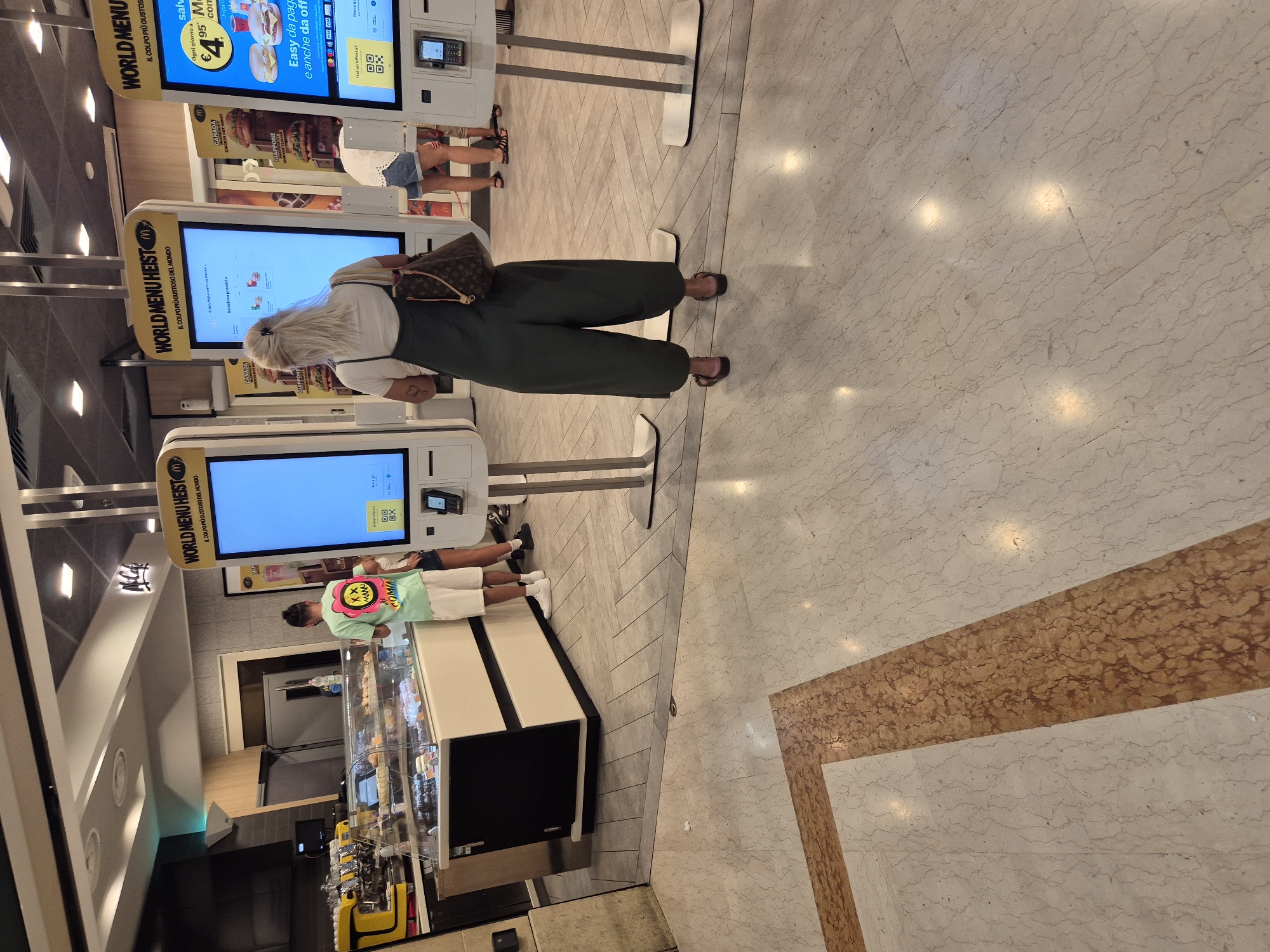 In a McDonald's restaurant in Taranto, Italy, several customers are interacting with self-service kiosks. In the foreground, a woman wearing a dark green jumpsuit and sandals stands with her back to the camera, looking at a digital menu. She has a Louis Vuitton handbag slung over her shoulder. To her left, a young man in a green t-shirt and white shorts is also looking at a kiosk. Further to the right, another person in a white shirt and denim shorts is partially visible, also near a kiosk. The environment is typical of a modern fast-food establishment, with sleek self-service terminals, brightly lit displays, and a clean, marble-patterned floor. Behind the kiosks, a glass display case showcases pastries and other food items, and a professional-looking coffee machine is visible. The overall scene suggests a typical day at a busy McDonald's location.