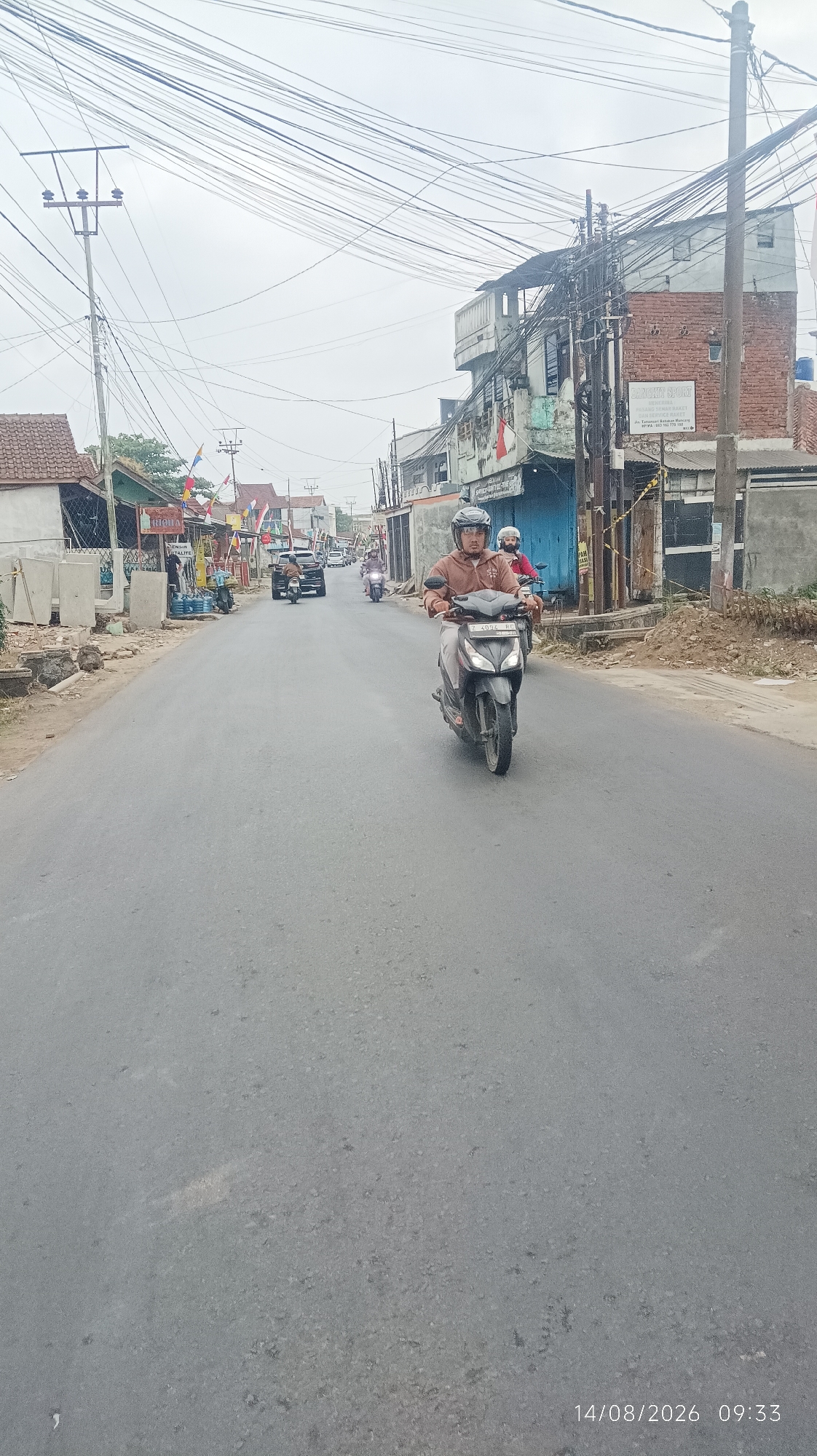 The image depicts a street scene in Tasikmalaya, Indonesia, on August 14, 2026, at 09:33. A person wearing a brown jacket and a helmet is riding a dark-colored scooter in the foreground, traveling away from the camera. Another person on a scooter is visible behind the first. The street is paved and appears to be a two-lane road with some traffic. Along the sides of the road, there are buildings, including a brick building with multiple stories and a sign that reads "AROMA SPORT." There are also utility poles with numerous overhead wires. The sky is overcast. The environment suggests a moderately developed urban or semi-urban area.