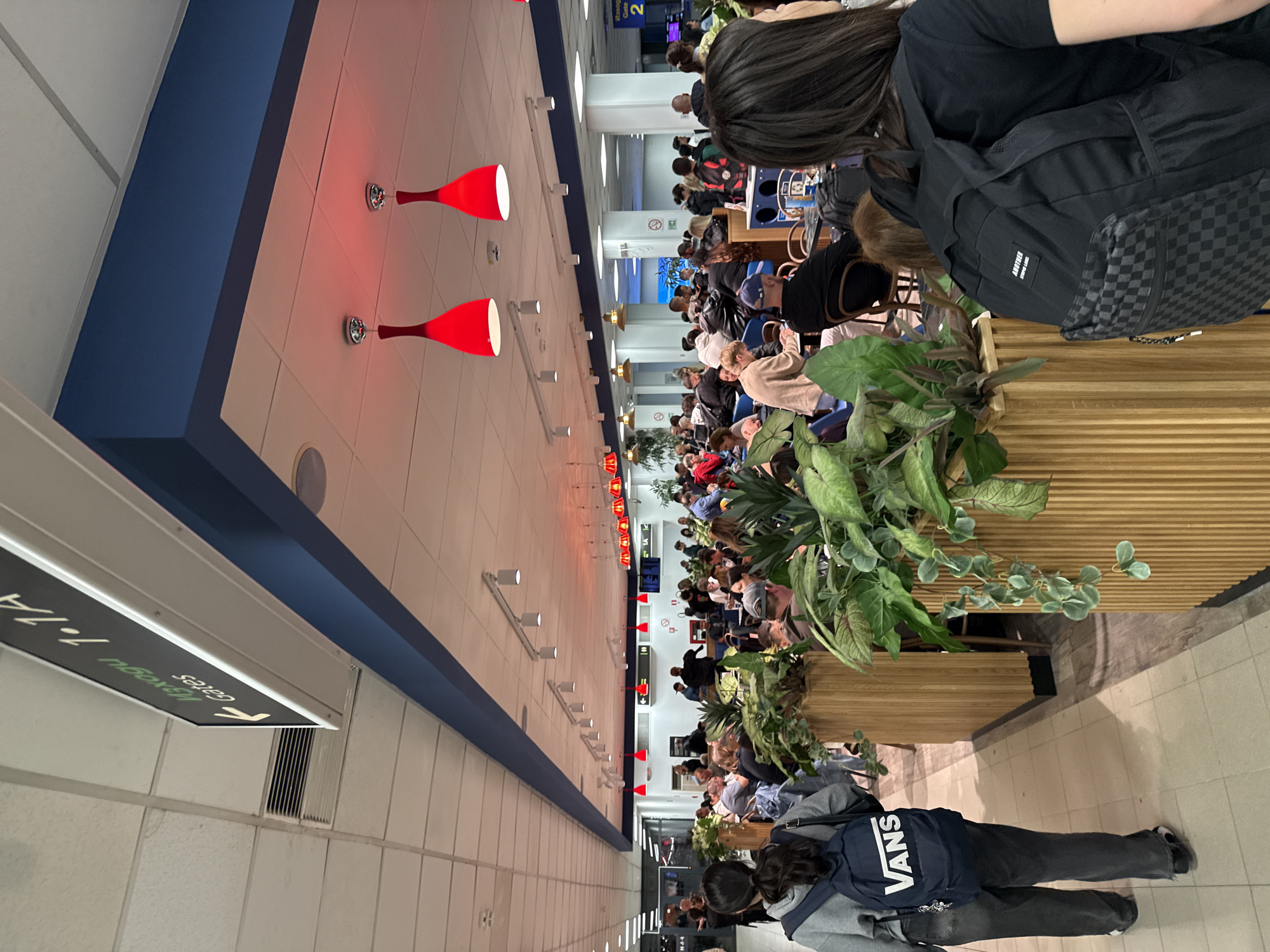 The image captures an indoor scene within what appears to be an airport terminal in Sofia, Bulgaria, oriented vertically but depicting a horizontal corridor. Numerous people are present, primarily seated in waiting areas, suggesting they are passengers. In the foreground, viewed from behind, a person with long dark hair wears a black top and a black backpack with a checkerboard pattern on the front pocket and a small white tag reading "ANOTHER STY-LE." Another person further back also has a blue backpack prominently displaying "VANS" in white lettering.
The activity is one of waiting or transit, common in an airport setting. Passengers are seen occupying rows of seats, some appearing to be looking at their phones or conversing. Large potted plants with green foliage are strategically placed, acting as decorative dividers between waiting areas and walkways.
Notable architectural details include a white ceiling with recessed lighting and several distinctive, red, cone-shaped light fixtures hanging down. A prominent blue trim runs along the top of a wall or structural beam, adding a splash of color to the otherwise neutral palette of the interior.
Visible text confirms the location type: a hanging sign in the foreground, which appears mirrored and upside down, shows the word "gates" with an arrow and the numerical range "7-11A." Another sign further in the background also indicates "gates 2." No specific time of day is discernible due to the indoor lighting. The general atmosphere seems calm, with people passively awaiting their departures or arrivals.