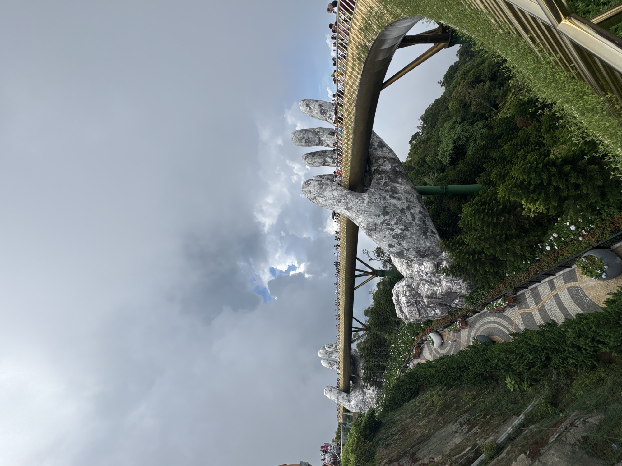 The image depicts the Golden Bridge (Cầu Vàng) held by giant stone hands in Bà Nà Hills near Đà Nẵng, Vietnam. A crowd of people is visible on the bridge. Lush greenery and ornate pathways surround the hand structures. The sky above is filled with dramatic, dark clouds. The bridge structure is supported by pillars.