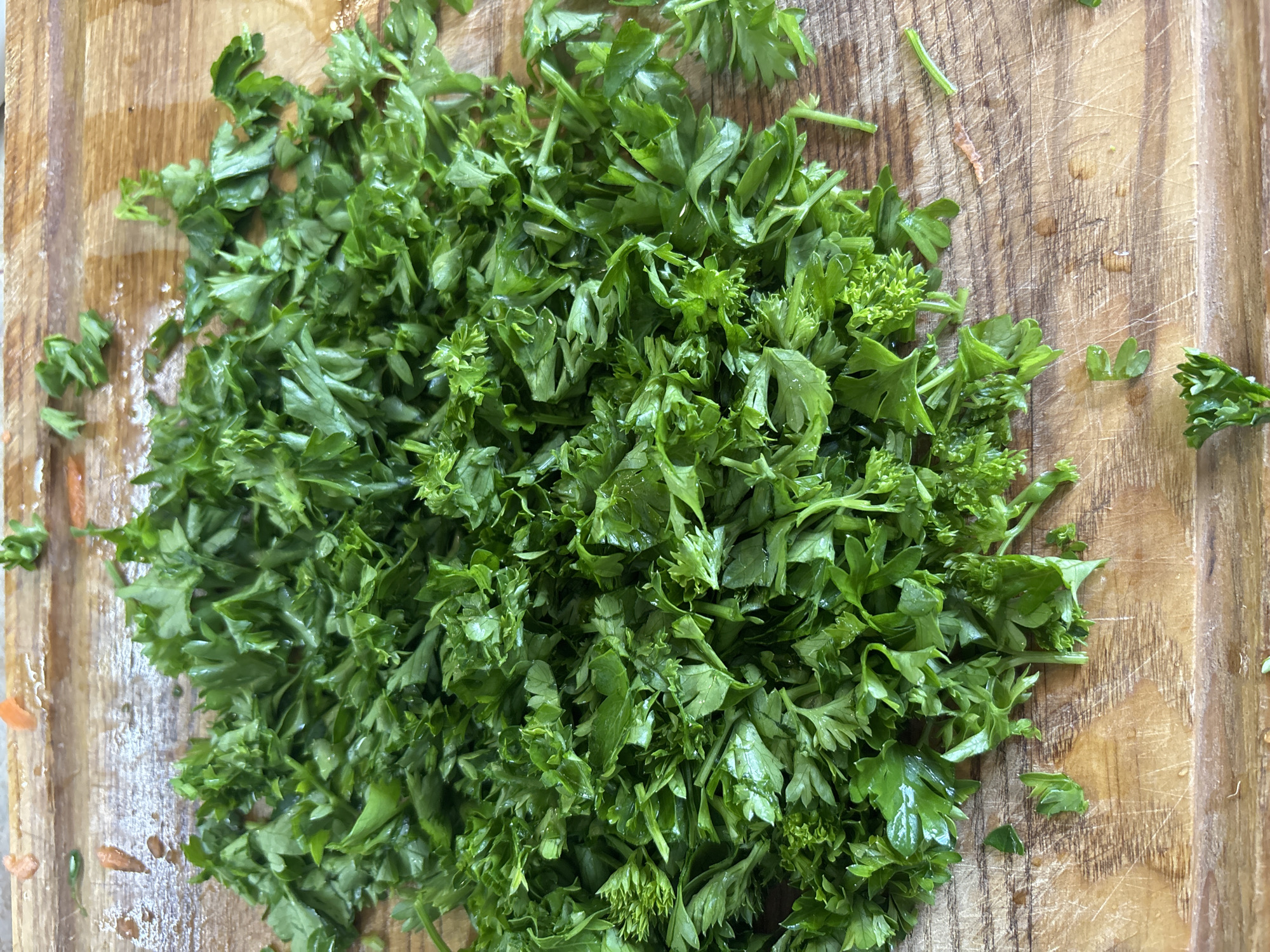 A pile of chopped fresh parsley sits on a wooden cutting board. The parsley is bright green and appears to be slightly wet, indicating it may have been recently washed. The cutting board has visible wood grain and a few faint scratches, suggesting it is well-used. There are also a few stray pieces of parsley scattered on the board, some of which are larger than the chopped pieces. The lighting suggests it is daytime, likely indoors, given the uniform illumination and lack of harsh shadows. The scene is simple and domestic, evoking a sense of food preparation. No people are visible in the frame. The location context of Bila Tserkva, Ukraine is noted but not visually discernible from the image itself. There is no visible text in the image.