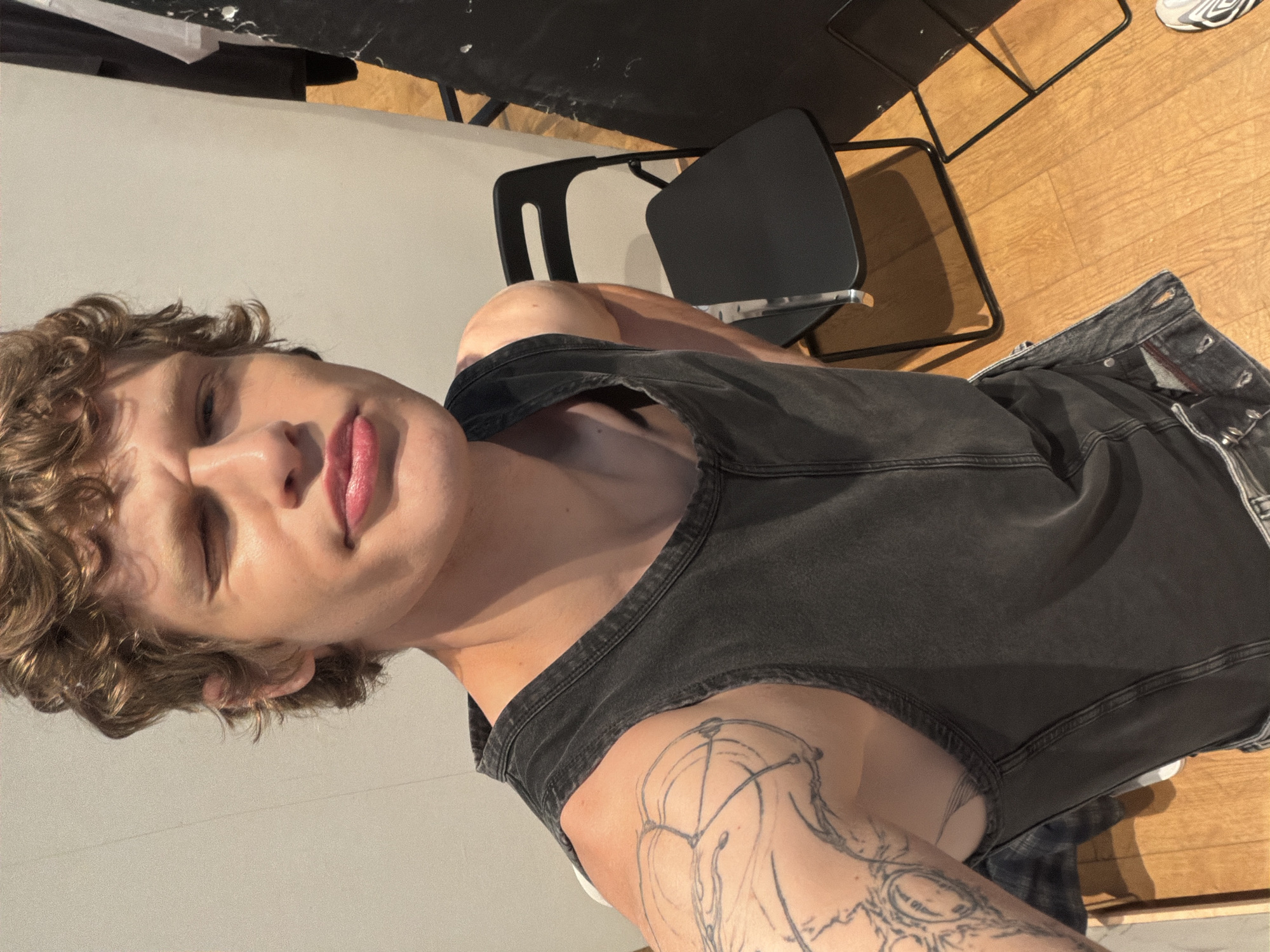 A light-skinned person with curly blonde hair, wearing a gray tank top and denim jeans, is angled towards the camera with their face at the top-left. The person's left eye is slightly closed. A tattoo is visible on their left shoulder. A black office chair is behind and to the right of the person. A wooden floor is at the right and top-right. A white wall is at the left. A white object is at the top-left. The location is Unknown, Unknown.