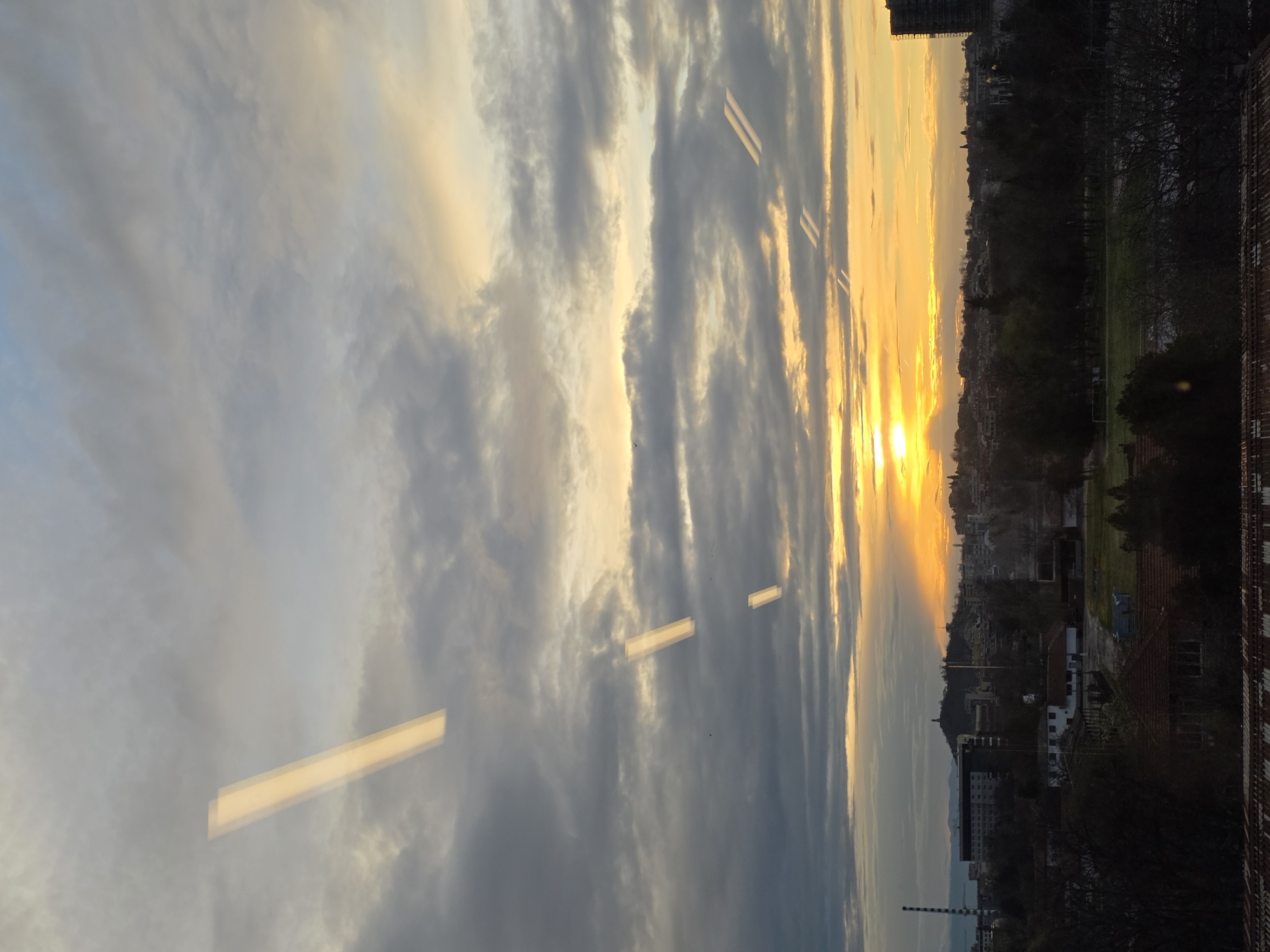 The image presents a view from a high vantage point, likely from within a building, overlooking a cityscape at sunset. The sky is filled with clouds illuminated by the golden light of the setting sun, creating a dramatic and colorful display. The sun's rays filter through the clouds, casting a warm glow over the urban landscape below.
The city itself is visible in silhouette against the bright sky. Buildings, trees, and possibly a park can be discerned in the middle ground. In the foreground, part of the building from which the photograph was taken is visible, including window reflections and possibly part of the building's exterior. The image is oriented sideways.
Based on the appearance of the cityscape, the presence of a hill with a monument on top and an industrial chimney on the left and the overall architectural style, the city is very likely Plovdiv, Bulgaria, and the hill is probably "Bunardzhik Tepe" (Liberators' Hill) with the Alyosha Monument visible on its summit. The industrial chimney is from Trakia Glass Bulgaria.