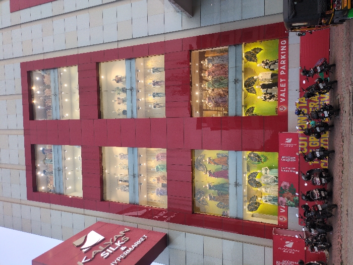 The image displays the exterior of the "Kalyan Silks & Hypermarket" building located in Ernakulam, India. The multi-story structure features a dominant red central facade, flanked by lighter, possibly off-white, vertical sections. The red facade is punctuated by six large, illuminated rectangular display windows arranged in a grid, each containing multiple mannequins dressed in traditional Indian garments such as sarees and dresses. The mannequins are positioned against varied backdrops, some with visible patterns. A red, angled sign on the left identifies the establishment as "Kalyan Silks & Hypermarket." At ground level, a row of approximately 15-20 motorbikes, primarily dark-colored, are parked in front of the building. The ground-level walls are red, displaying white text including "VALET PARKING" prominently on the right side and partial promotional text on the left. A dark-colored auto-rickshaw is partially visible on the far right of the frame. No human individuals are discernible within the visible scene.