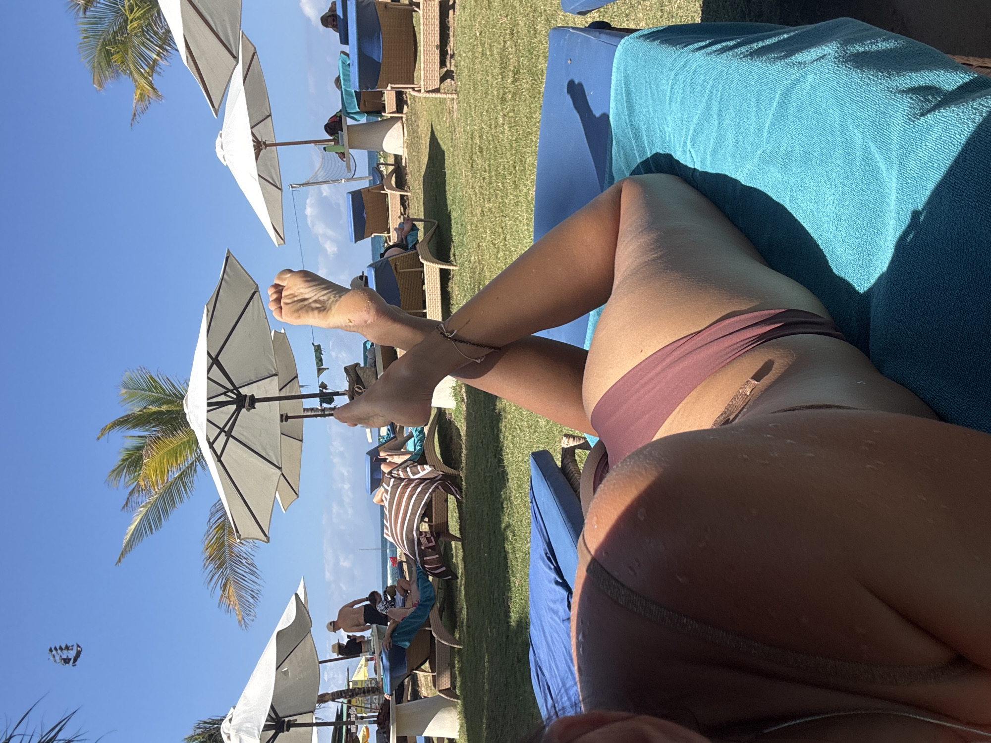 The image shows a person lying on a blue lounge chair on a grassy area, with a beach in the background. The person is wearing a bikini and their legs are crossed. There are several other lounge chairs and umbrellas on the beach, as well as some people walking around. In the sky, there is a kite shaped like a ship. The location is Kuta Selatan, Indonesia.