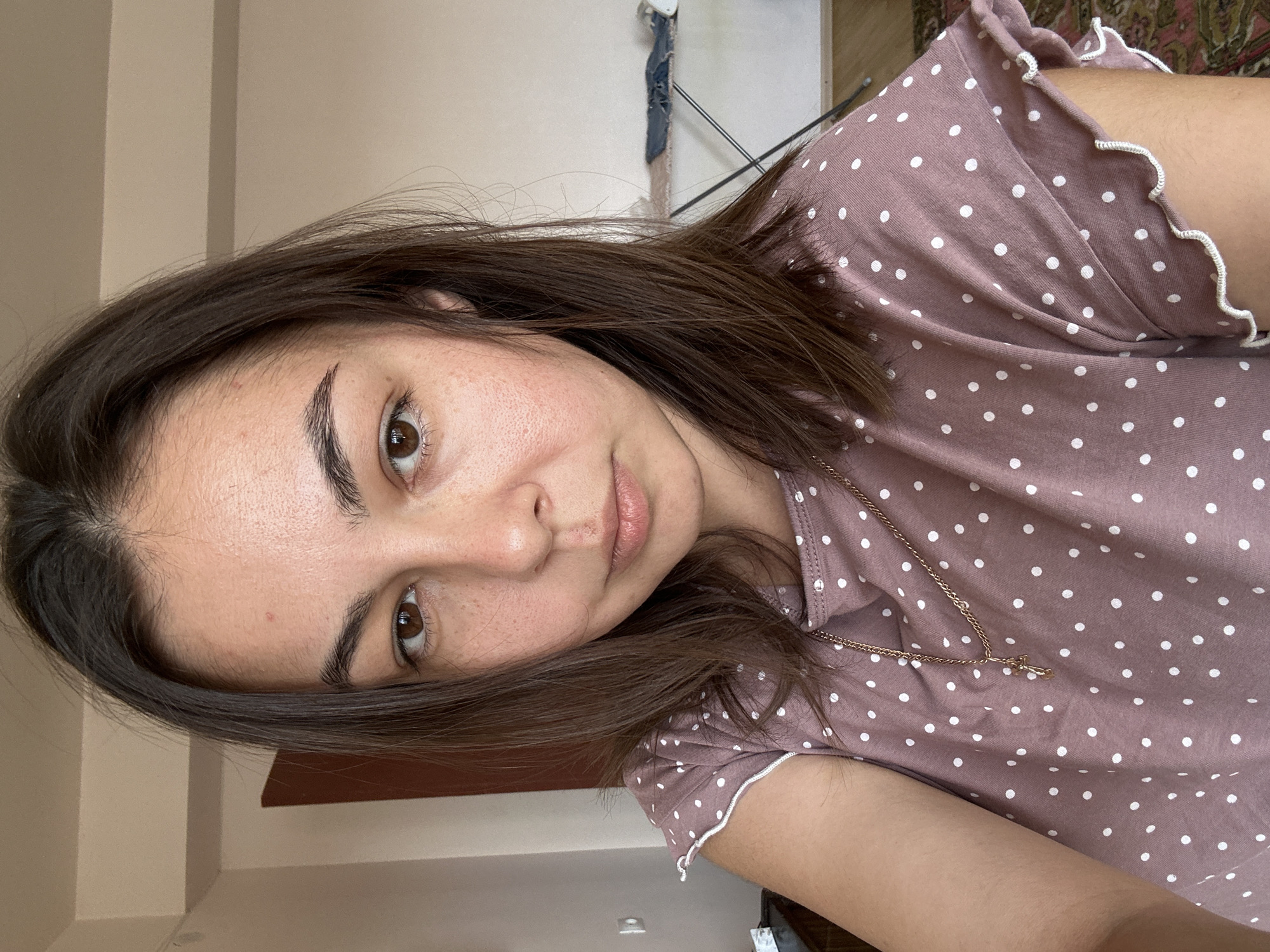 A young woman with shoulder-length brown hair and brown eyes is taking a selfie indoors. She is wearing a dusty rose-colored t-shirt with white polka dots and a delicate gold necklace. Her skin has a few blemishes and freckles. The background shows a light-colored wall and a door. To the right, an ironing board with some clothes is visible. The lighting appears to be natural, coming from a window to the right. The overall setting suggests a domestic environment. This image does not provide any information to suggest it is located in Gyumri, Armenia.