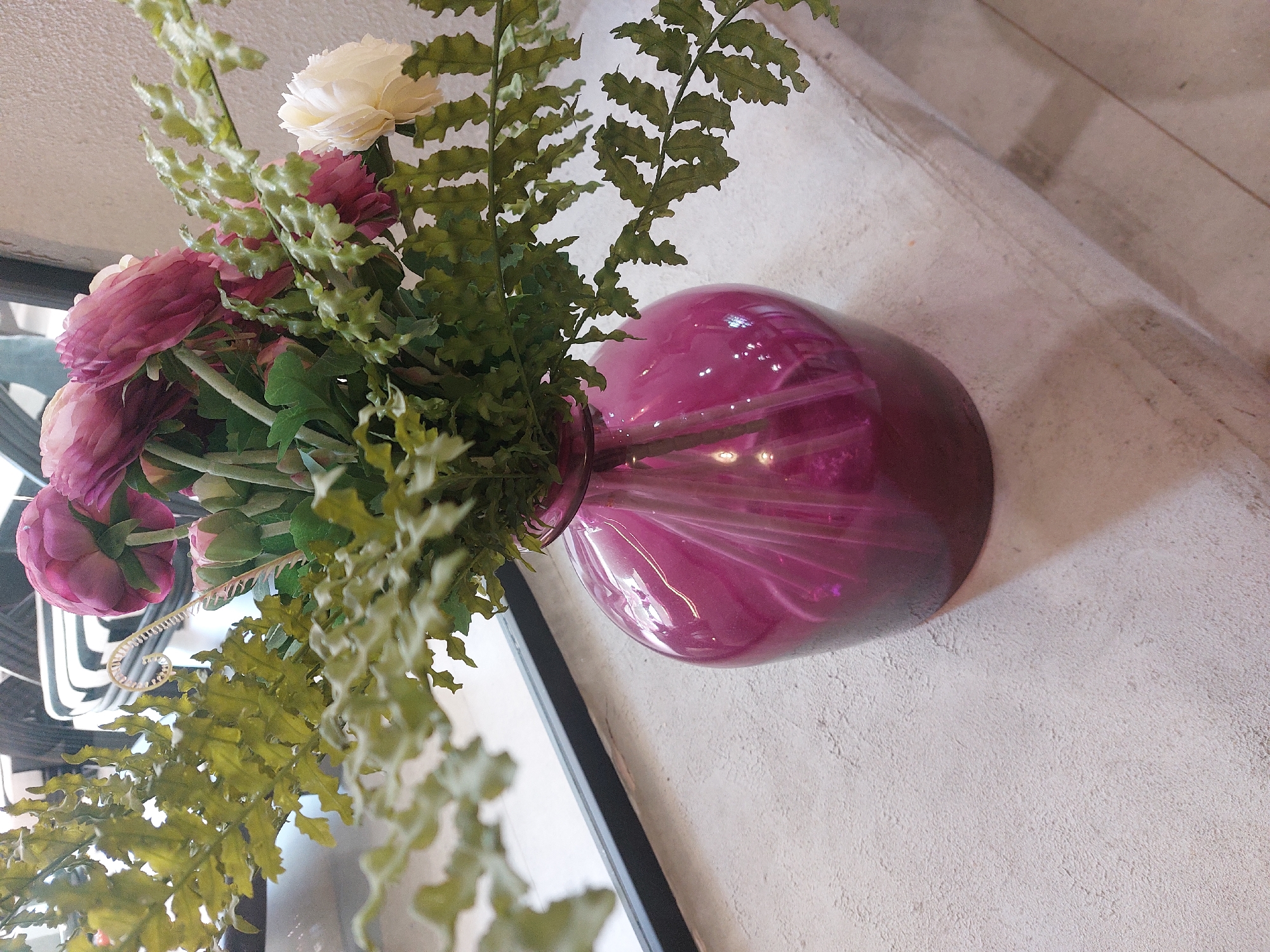 A close-up shot captures a bouquet of artificial flowers in a vibrant magenta glass vase. The flowers, a mix of deep pink and a single cream-colored bloom, are interspersed with delicate green foliage, resembling fern leaves. The vase is positioned on a textured, light-colored surface, possibly a windowsill or shelf, with a dark horizontal element visible behind it. The background is blurred, suggesting an indoor setting with natural light filtering in. The specific location in Kyzylorda, Kazakhstan, is not identifiable from the visual information provided.