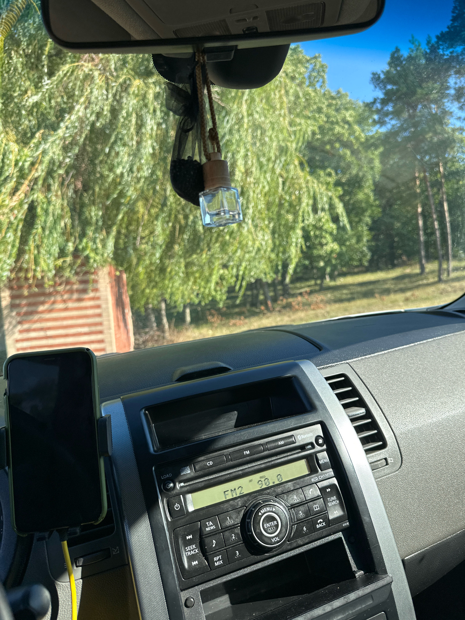 The image shows the interior of a car, looking forward through the windshield. A black smartphone is mounted on the dashboard to the left. To the right of the phone is a car stereo system. The stereo has a digital display showing "FM2 98.0". A car air freshener, a small glass bottle with a wooden cap, hangs from the rearview mirror. Outside the car, through the windshield, a sunlit scene of green trees and a blue sky is visible. The car appears to be parked on a road or a roadside, with the surrounding environment appearing rural or semi-rural, consistent with the outskirts of a settlement like Makiv, Ukraine. The lighting suggests it is daytime. There are no people visible in the image.
