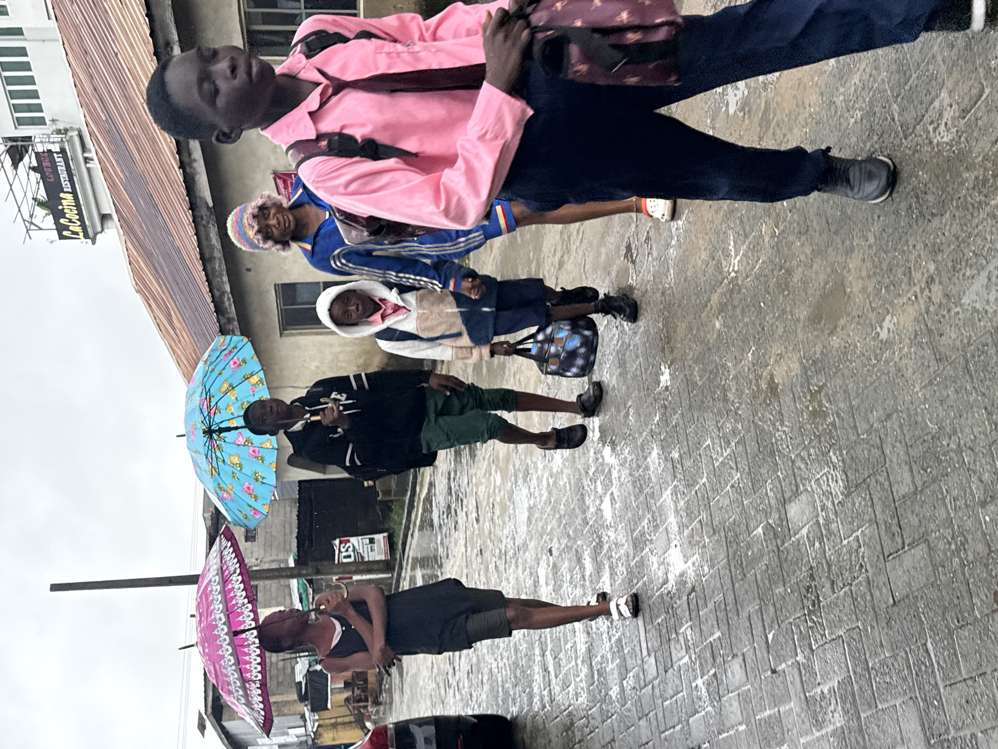 In Ado, Nigeria, under a cloudy sky and on a wet, brick-paved street, a group of children and an adult are walking. Two children are holding open umbrellas, one pink and patterned, the other blue with floral designs, to shield themselves from the rain. The child with the pink umbrella is on the far left, wearing a dark sleeveless dress over olive green shorts and white sandals. Next to them, a child in a black hooded jacket with white stripes and green shorts is also holding an umbrella. Further back and to the right, a young child in a white hooded jacket over a pink shirt and blue shorts walks with a patterned blue bag. Behind this child, an adult wearing a blue jacket and a colorful knitted hat is visible. On the far right, a child in a bright pink collared shirt and tie, navy pants, and dark sneakers walks forward, carrying a backpack with a patterned strap. The street reflects the overcast sky and the wetness from the rain. Buildings line the street, one with a sign reading "LaCocina Restaurant."