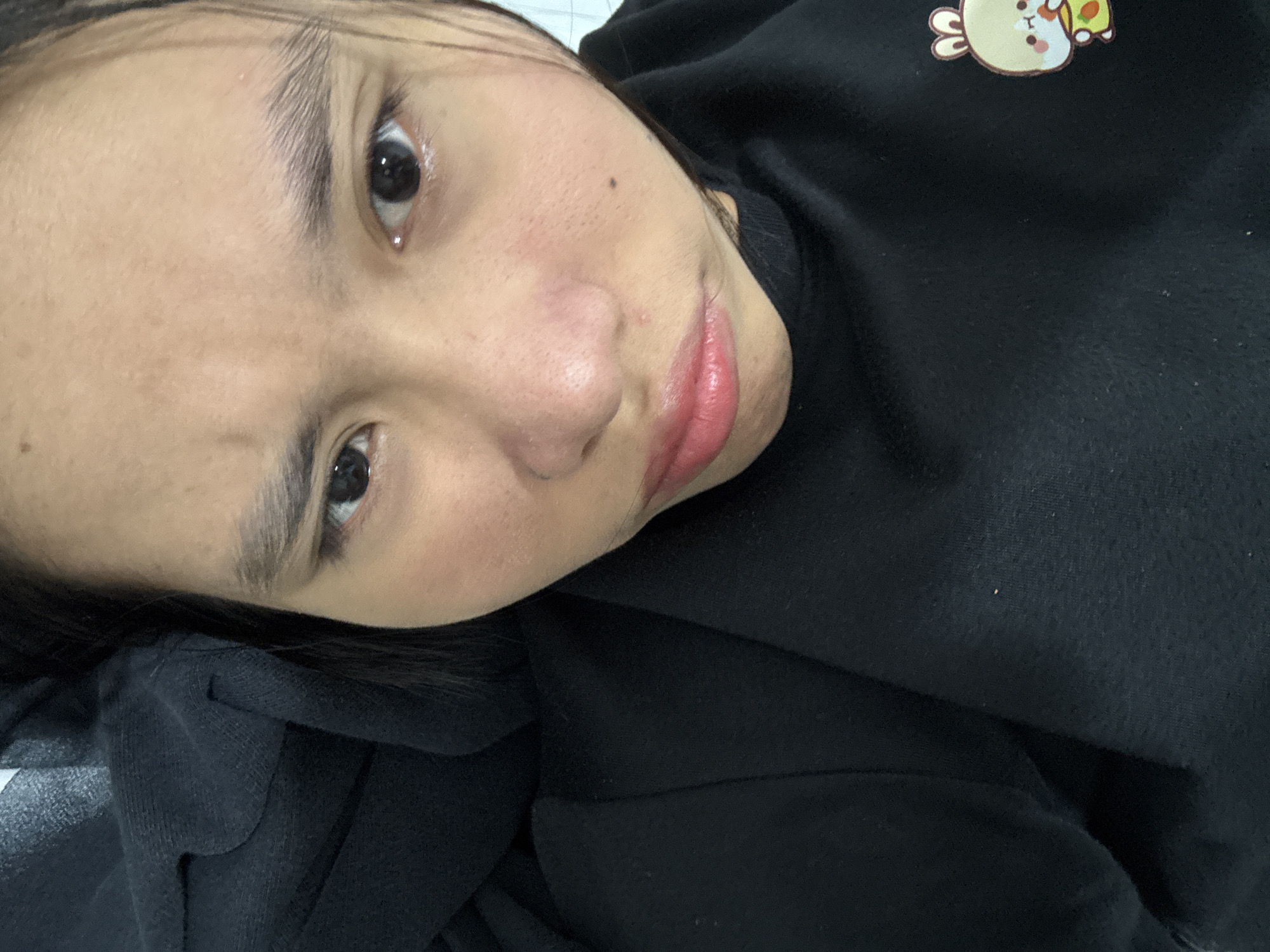 The image is a close-up selfie of a young East Asian woman, taken from a tilted perspective. She is lying down, resting her head on dark fabric, likely clothing. Her facial features are prominent: dark, expressive eyes, arched eyebrows, a small nose, and full lips with a pinkish hue. Her skin appears smooth with a few subtle freckles visible. She is wearing a black garment with a small, light-colored cartoon graphic on the upper right portion of the fabric, depicting a round character with ears and holding a green object. The background is indistinct, suggesting an indoor setting. The lighting appears to be ambient, casting soft shadows. There are no other people or significant objects in the frame. The image does not contain any recognizable landmarks or indicators of a specific location within South Jakarta, Indonesia.