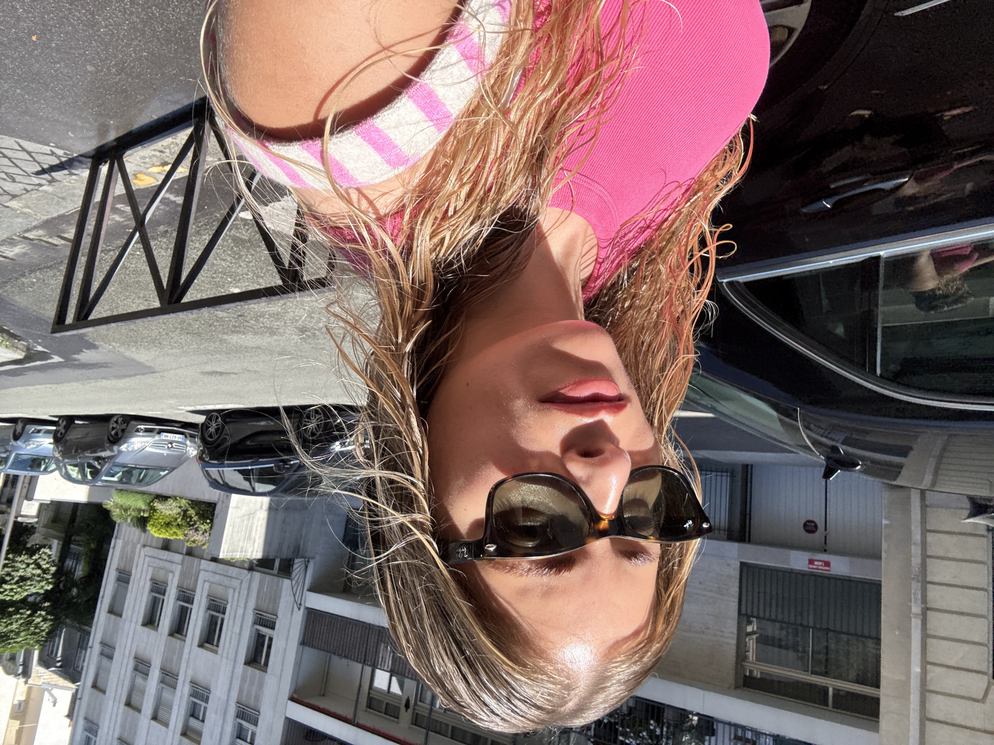 A woman with wet blonde hair is standing on a street in Paris, France. She is wearing sunglasses and a pink top. A black car is parked next to her. In the background, there are buildings and other cars parked on the street. The image is slightly rotated.