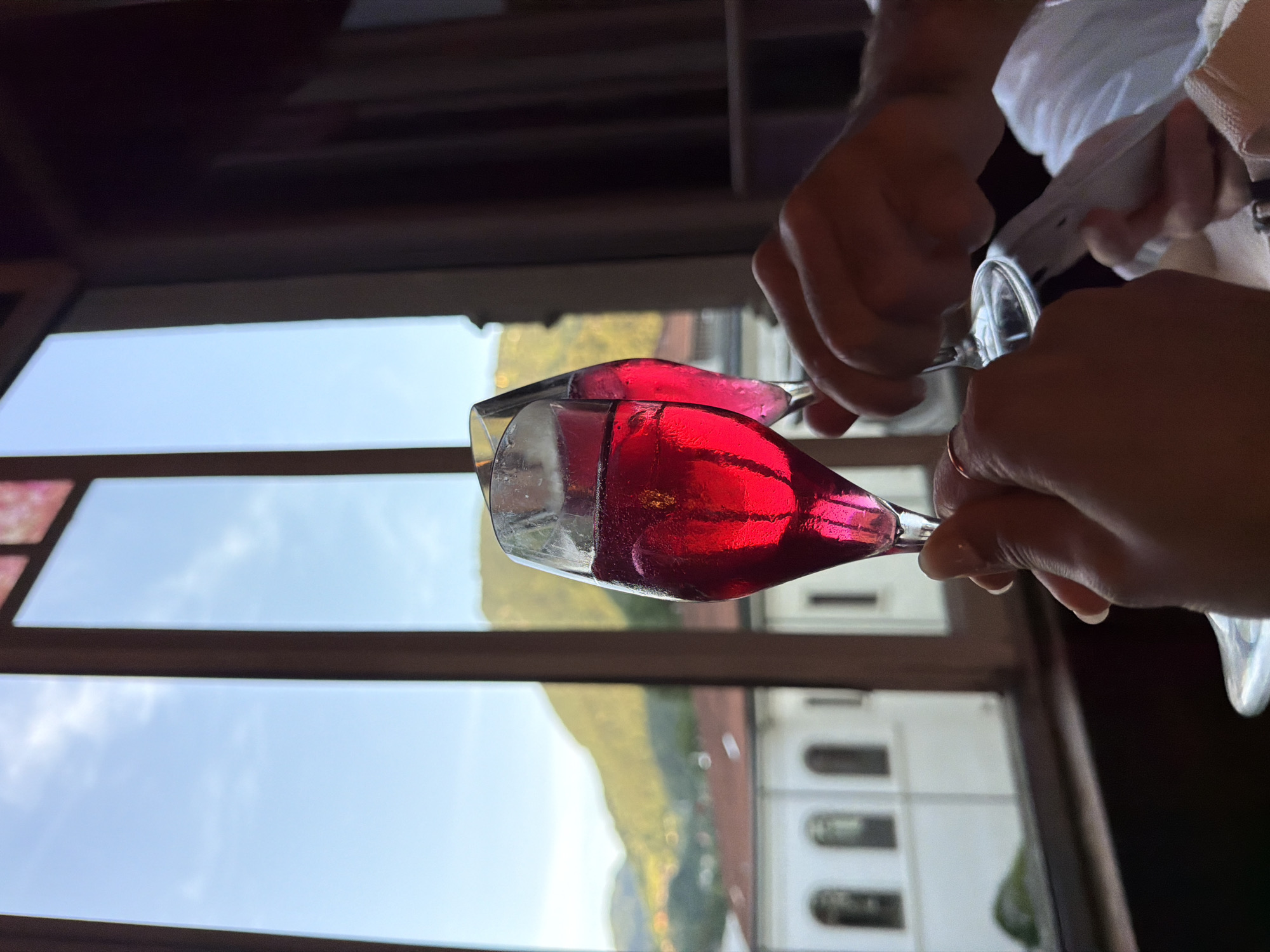 Two champagne flutes filled with a vibrant red liquid are being held up in a toast, their contents catching the light. In the background, through a large window, a scenic view of San Pellegrino Terme, Italy, is visible. The window frames rolling green hills dotted with buildings, and a bright blue sky with sparse clouds. The overall impression is one of celebration and enjoyment, possibly at a restaurant or a similar establishment in the Italian town.