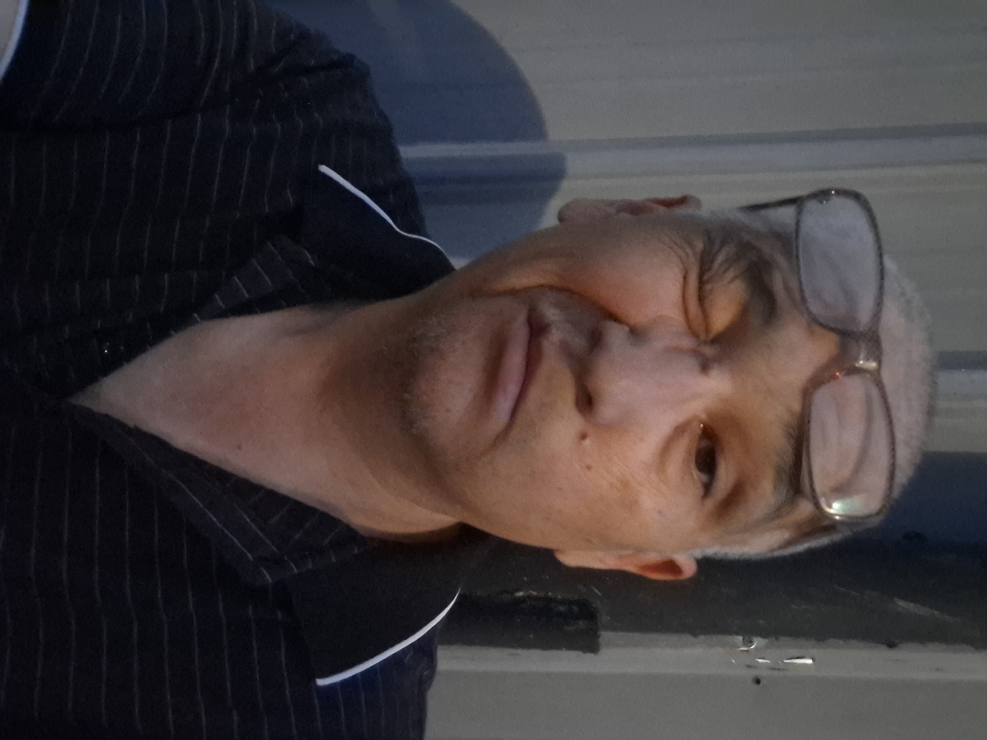A close-up, medium shot, chest-up photograph shows a middle-aged man of indigenous descent, with light skin, balding grey hair, and a light stubble. He is wearing glasses that are slightly askew on his head, and he is squinting his right eye. His left eye is open and looking directly at the camera. He is wearing a dark, pinstriped polo shirt. The man is positioned in front of a ribbed metal wall, which appears to be part of an industrial building. To his right, there is a dark, cylindrical object, possibly a pipe or pillar, and a vertical line of reflective material, suggesting a seam or light source. The lighting is somewhat dim, with a shadow cast on the wall behind him. The image was captured outdoors, as indicated by the natural light and the texture of the surroundings, which could be found in an industrial area or an alleyway within the city of Almaty, Kazakhstan, potentially near the industrial zones such as those found in the southern parts of the city.