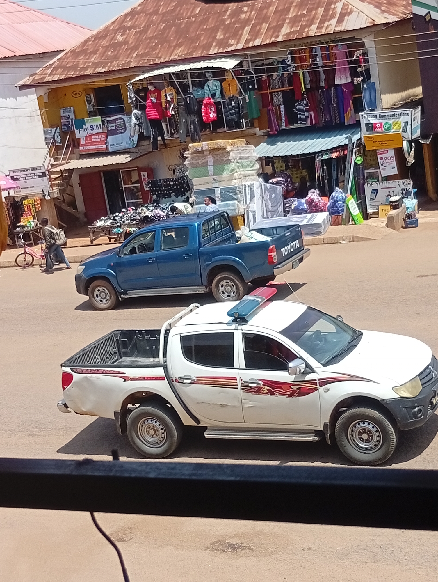 In Bukuru, Nigeria, two white pickup trucks are parked on a dusty road. The truck in the foreground has a light bar on its roof, and the truck behind it is blue and has "TOYOTA" written on its tailgate. The road is lined with shops selling clothes and other goods. In the background, a man is walking with a bicycle. The scene appears to be a busy market area, with several people visible in and around the shops.