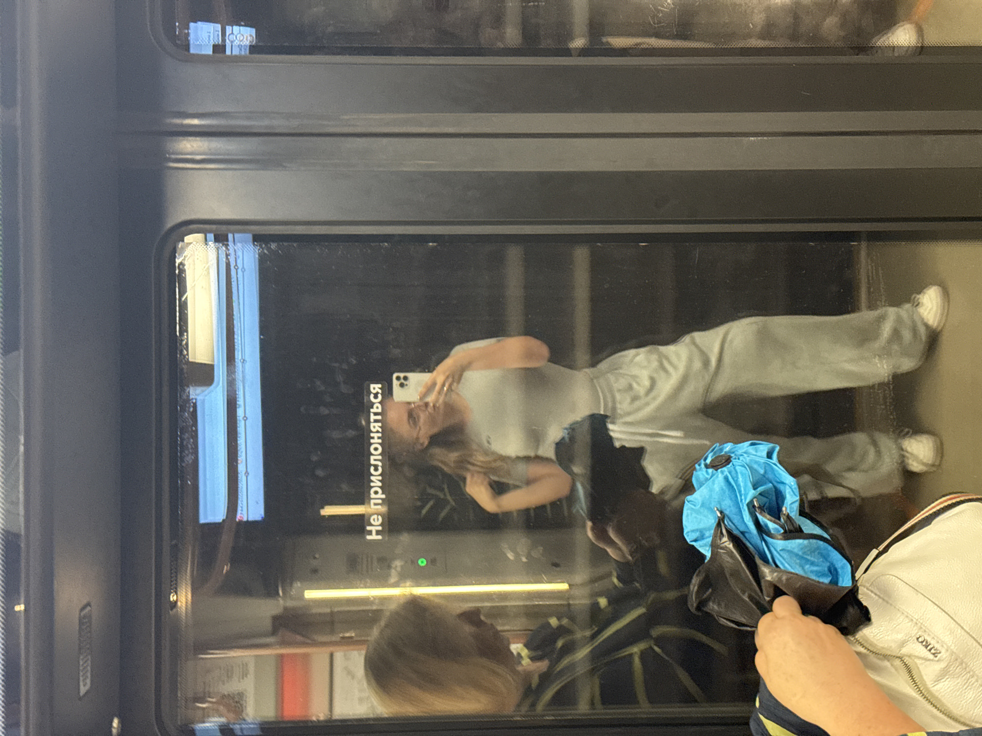 In Moscow, Russia, a woman is reflected in the doors of a metro train. She is taking a selfie with her phone, wearing a grey t-shirt and light-colored track pants. She has her right hand on her chest, and her left hand is not visible. In the reflection, another woman with blonde hair is visible to the left, wearing a striped shirt and holding a blue and black object. The metro car interior is dimly lit with yellow and white lights. A sign with Cyrillic text that translates to "Do not lean" is visible on the door.