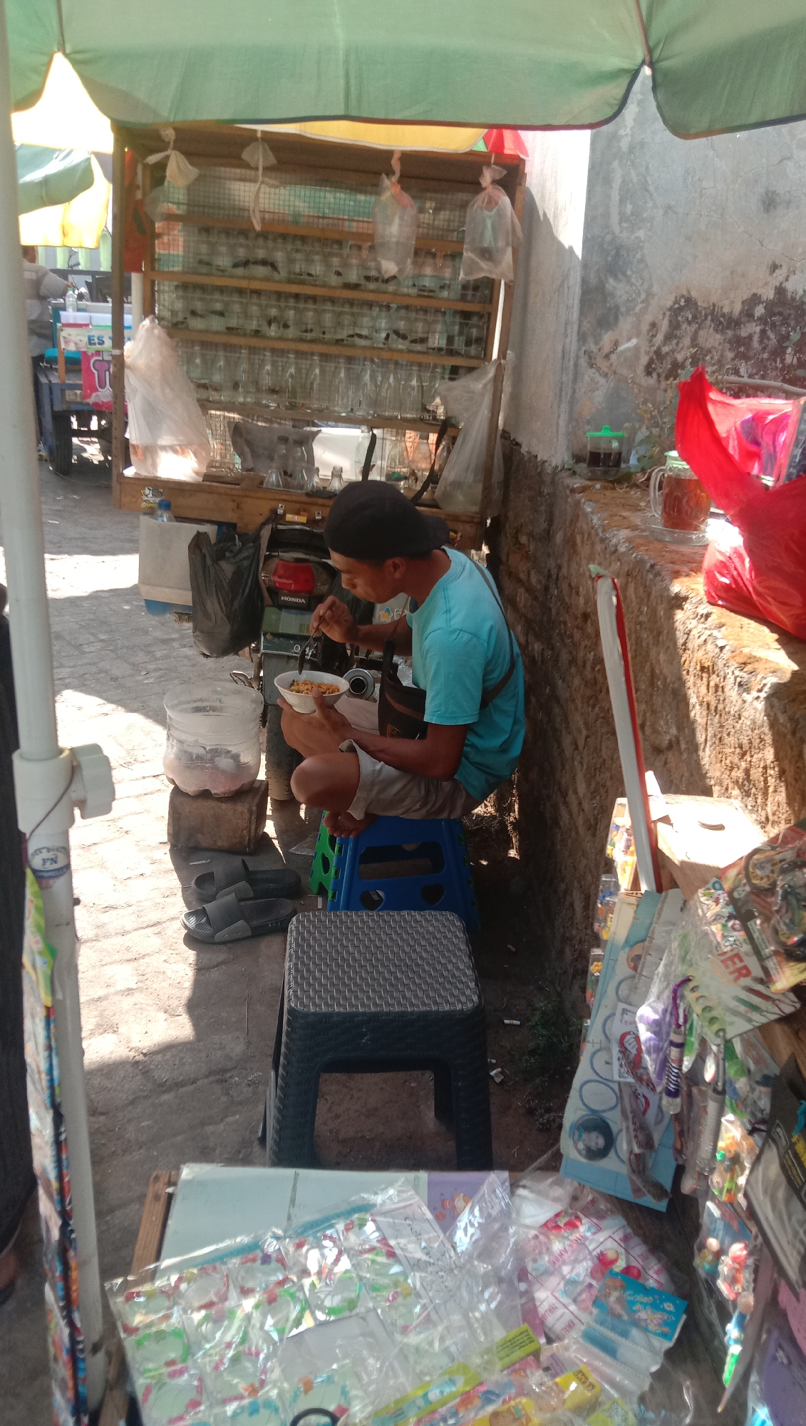 The image depicts an outdoor scene, likely a street vendor stall in Karang Anyar, Indonesia. A young man wearing a blue t-shirt and a black cap is seated on a blue plastic stool, eating from a bowl with a spoon. He is positioned next to a cart laden with rows of glass jars, each containing what appear to be fish or aquatic creatures. A large clear plastic container filled with water and ice is placed on the ground near him. Adjacent to this, a pair of grey flip-flops are visible. Behind the man, a motorcycle is partially visible, with the "HONDA" logo clearly discernible. The stall itself is covered by a green and yellow awning. In the foreground and to the right, a variety of items are displayed, including packaged goods, possibly toys or snacks, suggesting the vendor sells multiple types of merchandise. The ground appears to be paved with interlocking bricks. The overall environment suggests a warm, sunny day.