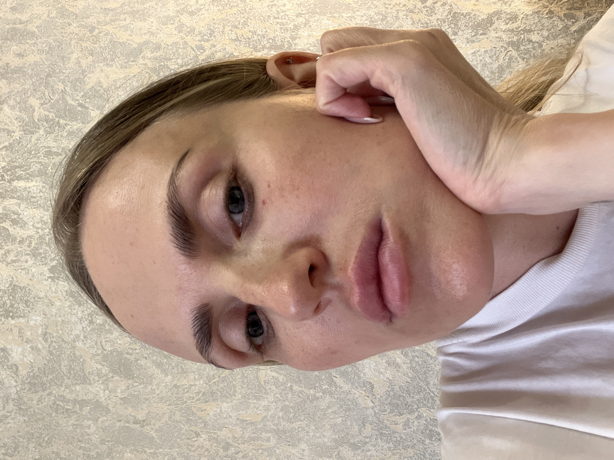 A close-up, head-and-shoulders shot of a woman rests her chin on her hand, her elbow bent and forearm visible. She wears a white t-shirt. Her blonde hair is pulled back. The background is a textured wall in shades of beige and gray.