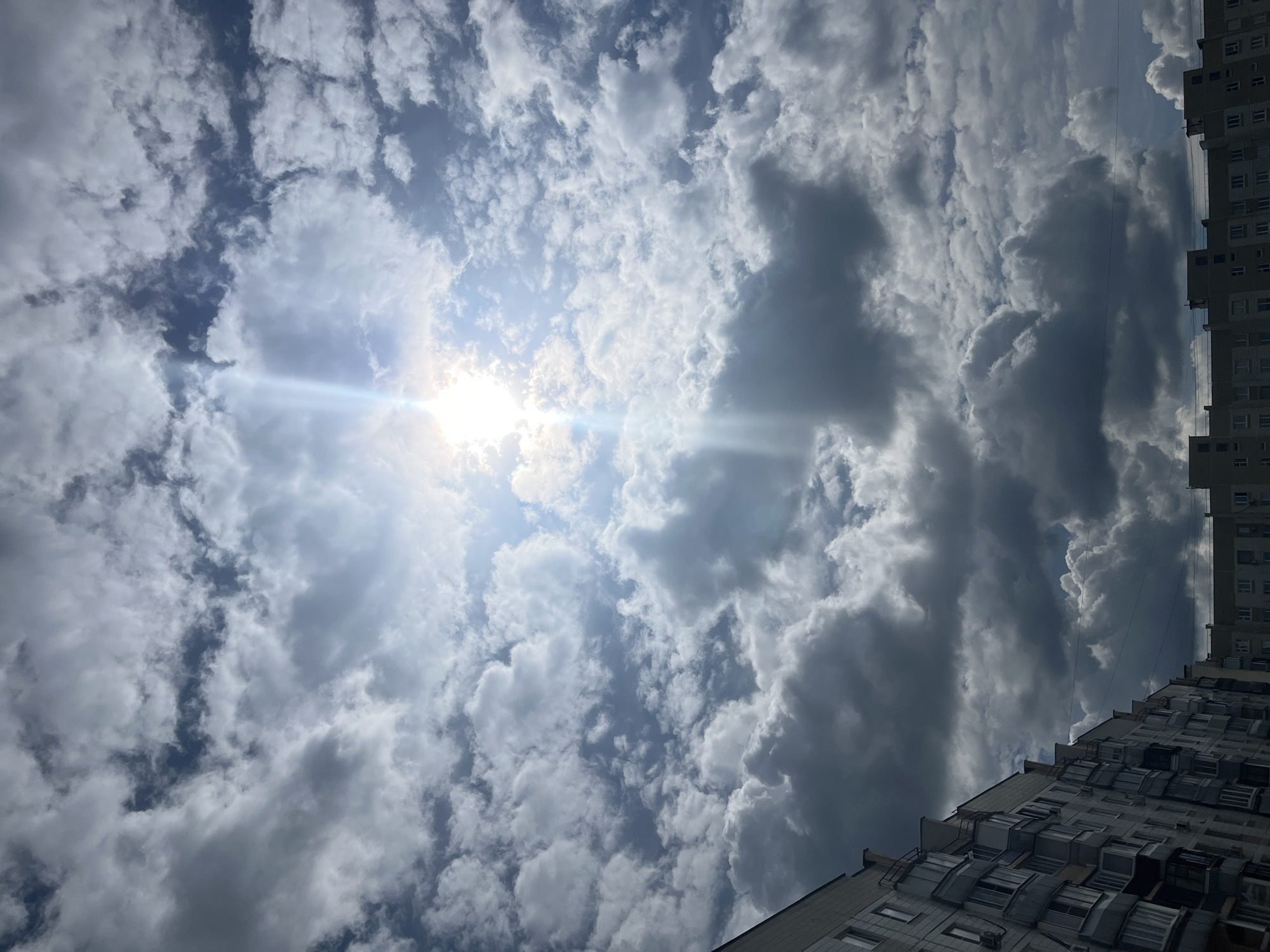 The image shows a view of a cloudy sky with the sun shining through, creating a bright sunburst effect. In the lower left corner, the side of a multi-story apartment building is visible, with windows and balconies. The sky is filled with a mix of white and gray clouds, some of which are dark and appear heavy. The sun is located near the top center of the image. The scene appears to be an urban environment. The question asks for the location in Moscow, Russia; however, the image itself does not provide any explicit information to confirm this location. Therefore, I cannot confirm the city or country.
